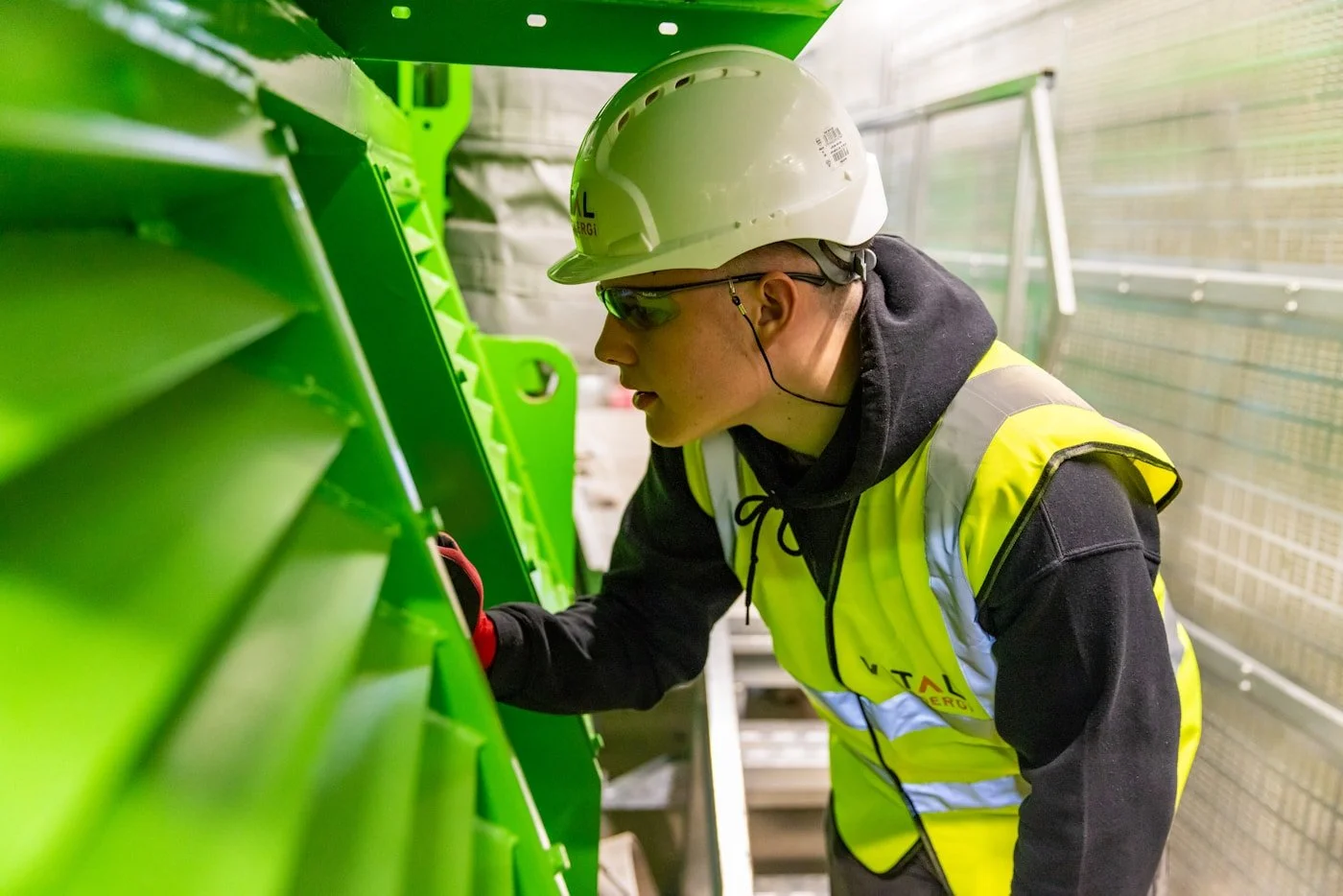 A worker wearing a white hard hat, safety glasses, a yellow safety vest, and black hoodie is inspecting or repairing green electrical or mechanical equipment inside an industrial or power plant setting.