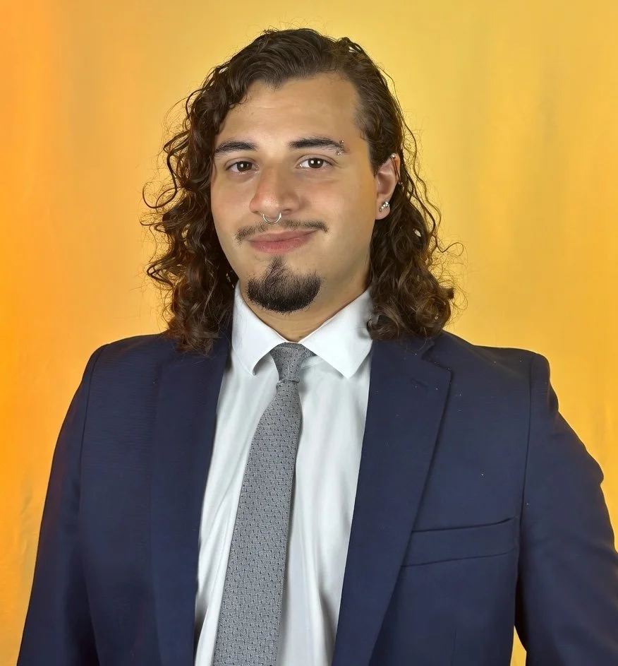 A young man with shoulder-length curly hair, a mustache, and beard, wearing a dark suit, white shirt, and gray tie, standing against a yellow background. Kevin Anthony Garcia