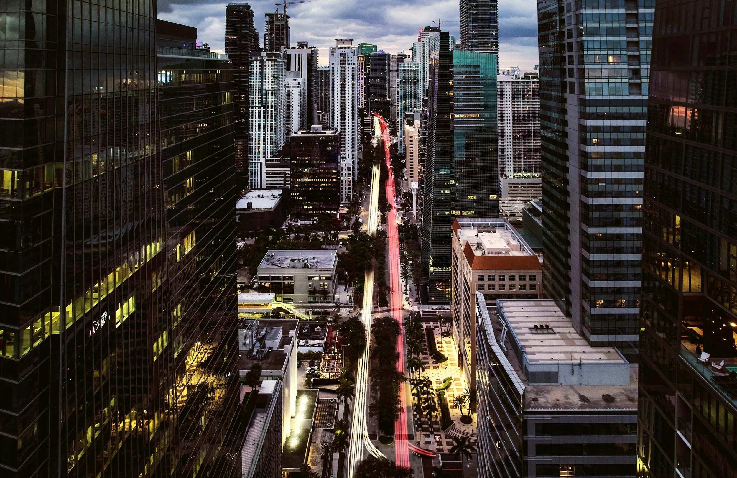 Nighttime aerial view of downtown city with light trails from moving vehicles on a busy street, high-rise buildings, and a cloudy sky.