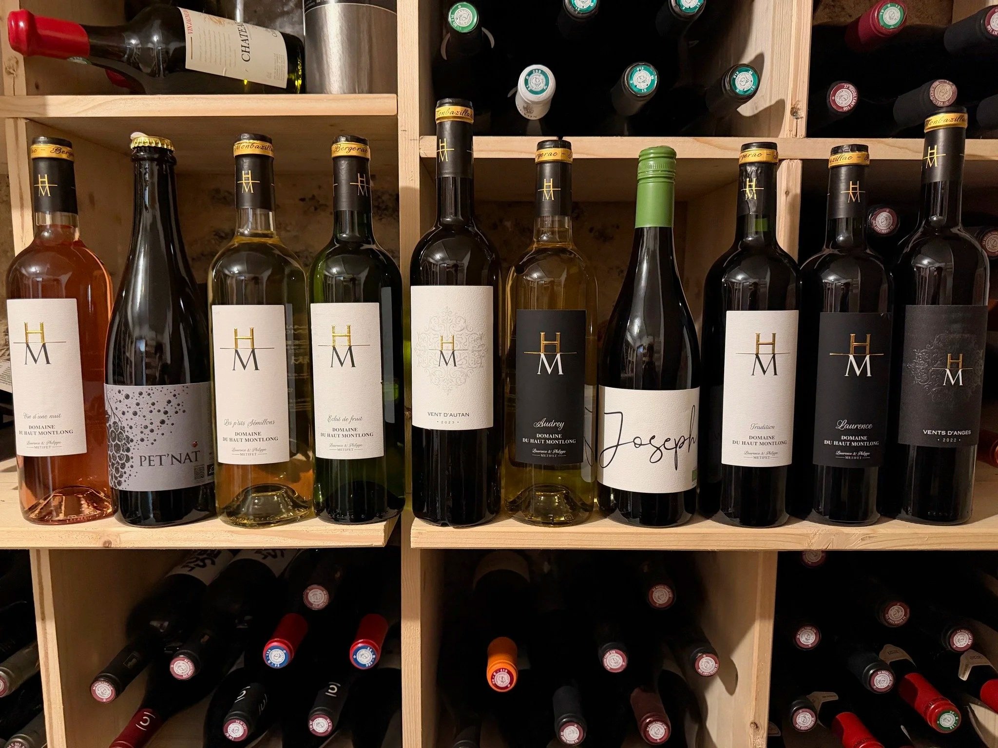 Une étagère en bois contenant plusieurs bouteilles de vin de différents types et étiquettes, avec d'autres bouteilles de vin empilées en dessous dans une cave à vin.