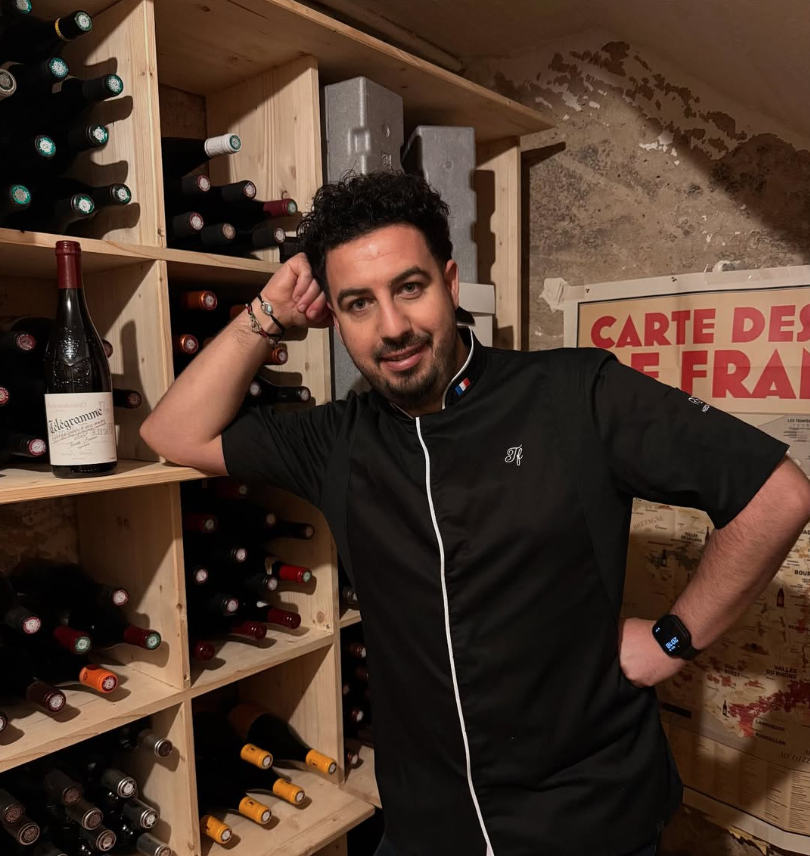 Un homme en chef noir pose dans une cave à vin, entouré de bouteilles de vin sur des étagères en bois.