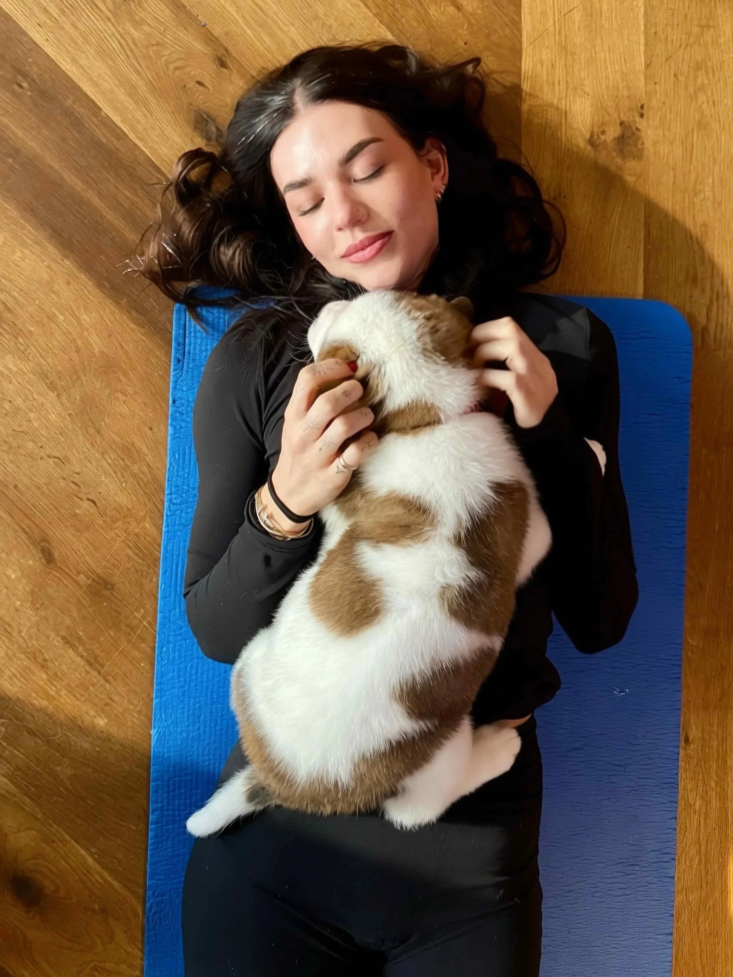 A woman with dark hair lying on a blue yoga mat on a wooden floor, gently cuddling a small puppy.