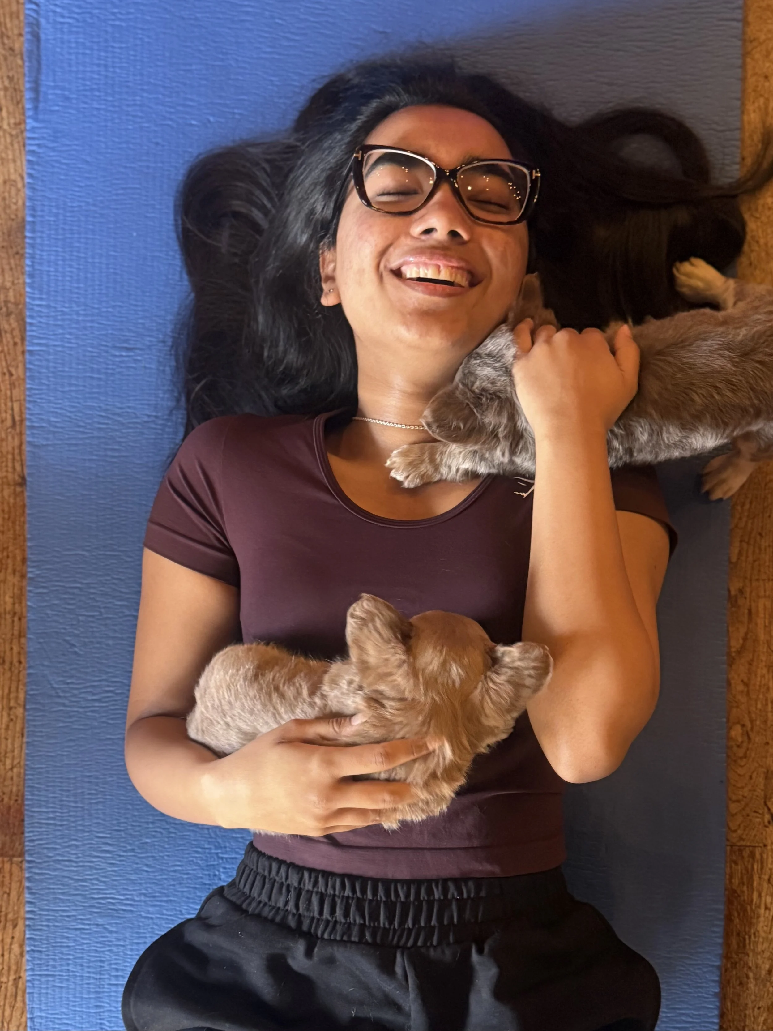 A woman lying on a blue yoga mat, smiling with eyes closed, holding two small puppies, one on her chest and one in her hand.