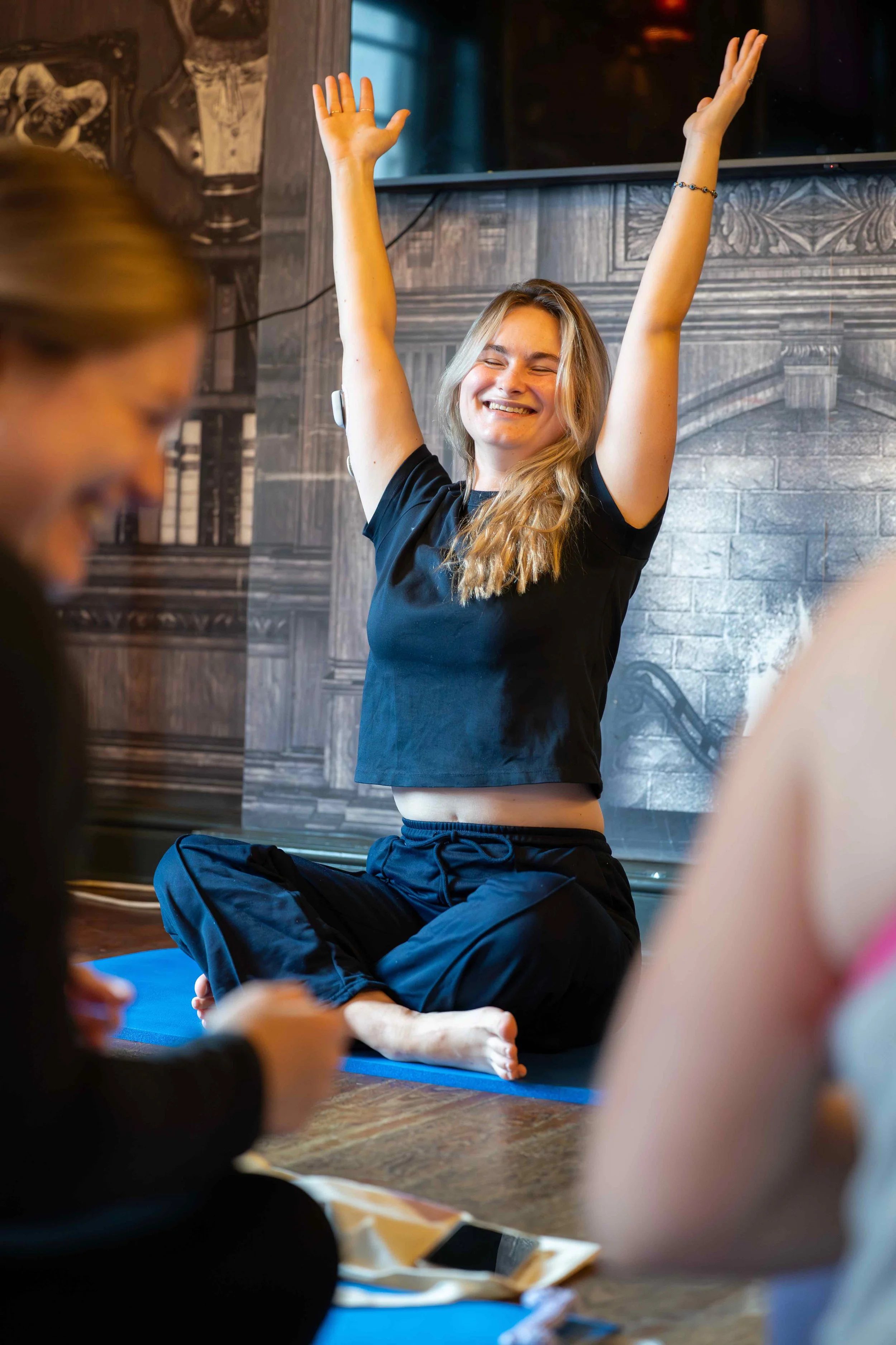 A woman sitting cross-legged on a yoga mat with arms raised and eyes closed, smiling.