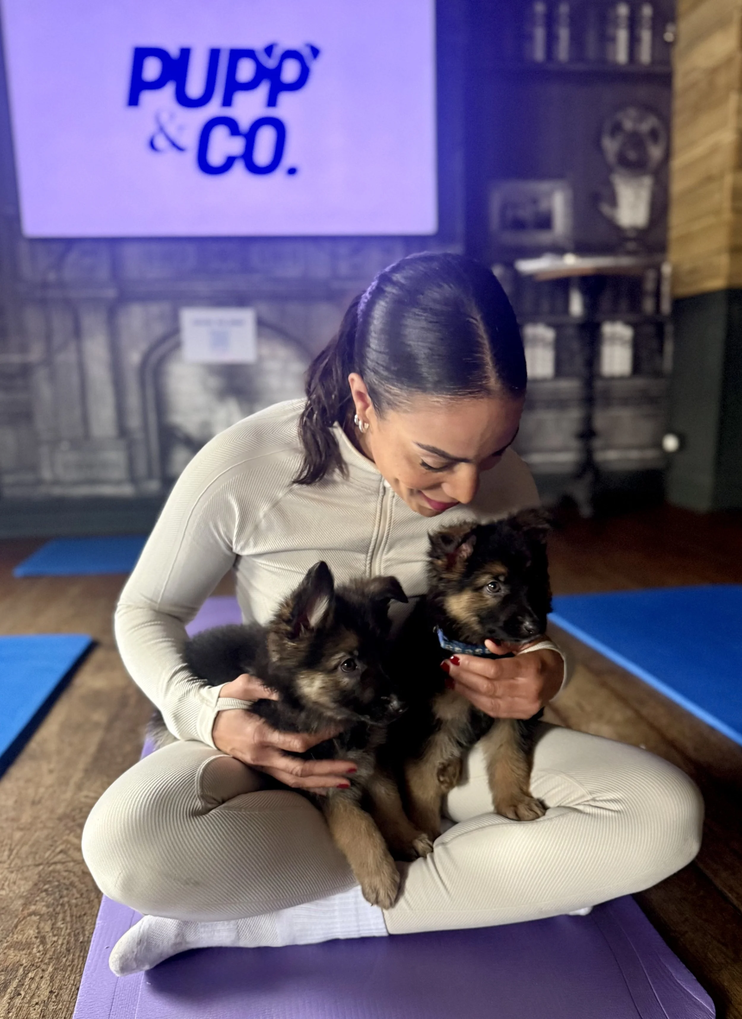 A woman sitting cross-legged on a purple yoga mat, holding two German Shepherd puppies in her arms. A large sign saying Pupp & Co is behind her.