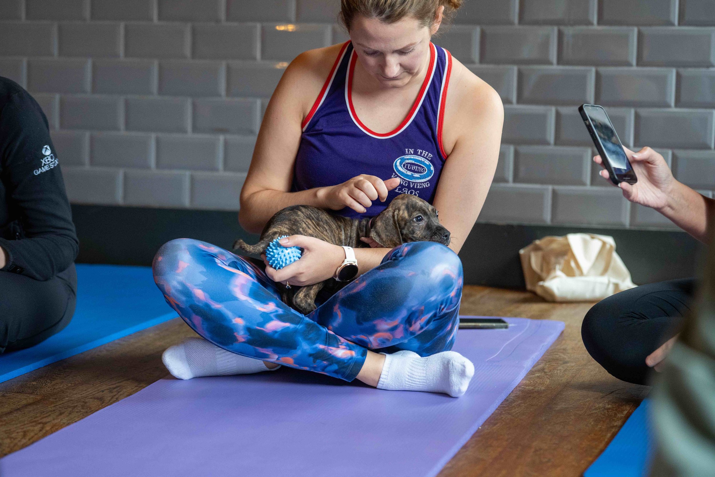 A woman in a fitness outfit holding a small puppy on her lap during a puppy yoga session. She is sitting cross-legged on a yoga mat, petting the puppy with one hand while holding a blue dog toy in the other.