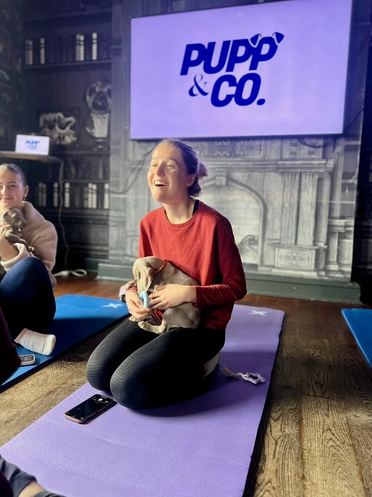 A woman with a puppy on her lap during a puppy yoga class. She is kneeling on a purple yoga mat in a studio with a large screen behind her displaying 'PUPP & CO.'