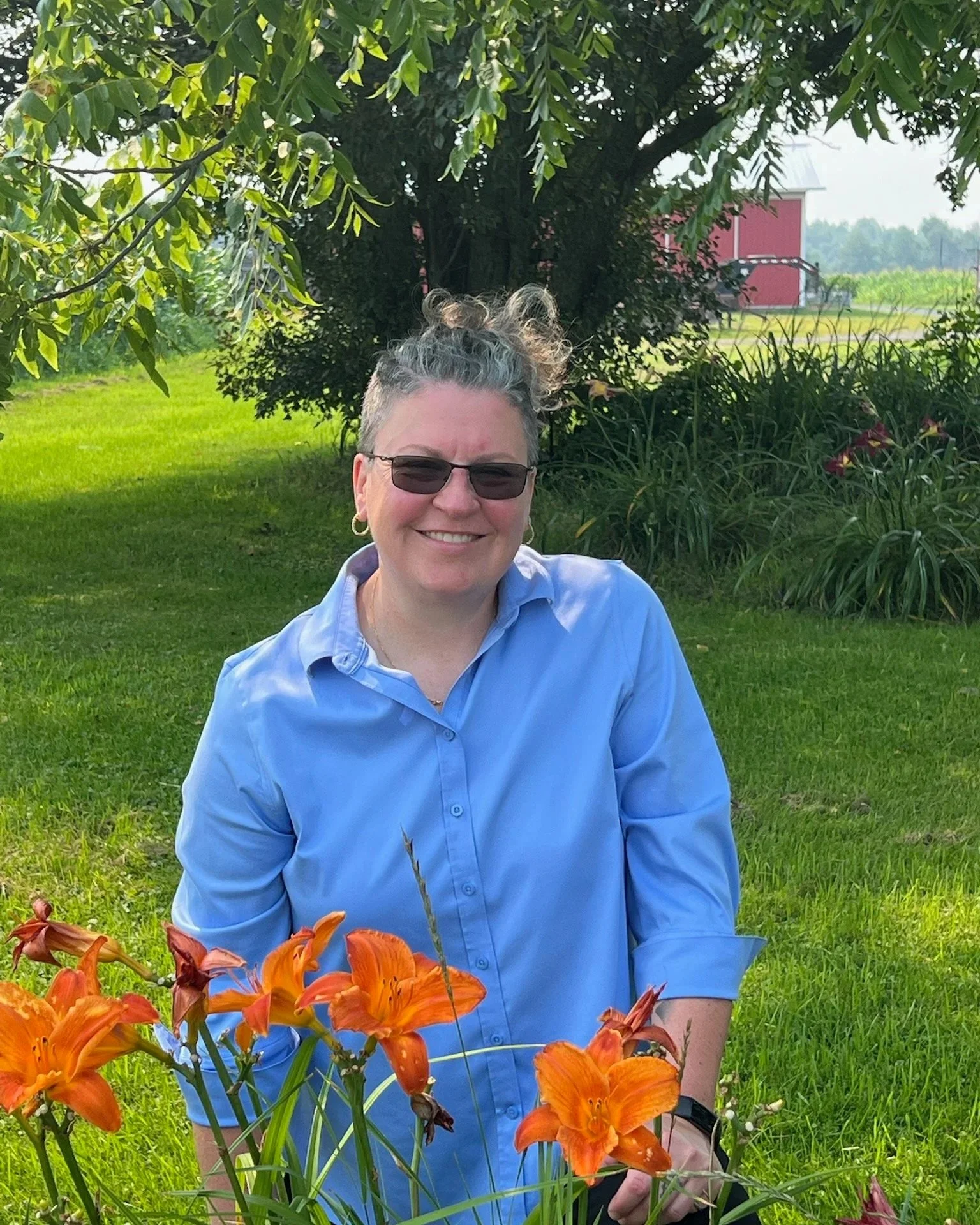 Angie Dittrich smiling, kneeling in a garden with orange daylilies, a red barn, and trees.