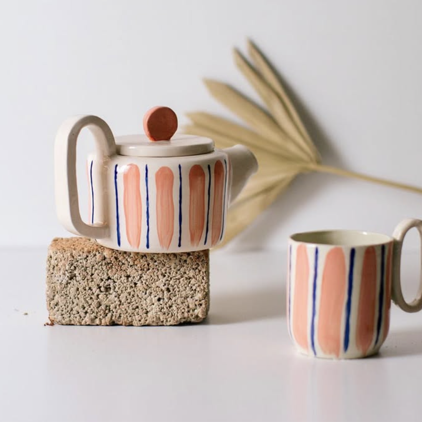 A decorative ceramic teapot with vertical orange, pink, and blue stripes, placed on a textured brick, with a matching mug nearby. A dried palm leaf in the background adds an artistic touch.