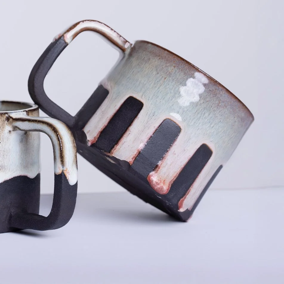 Close-up of two ceramic mugs with black and white glazed patterns, one mug leaning on the other, against a plain background.