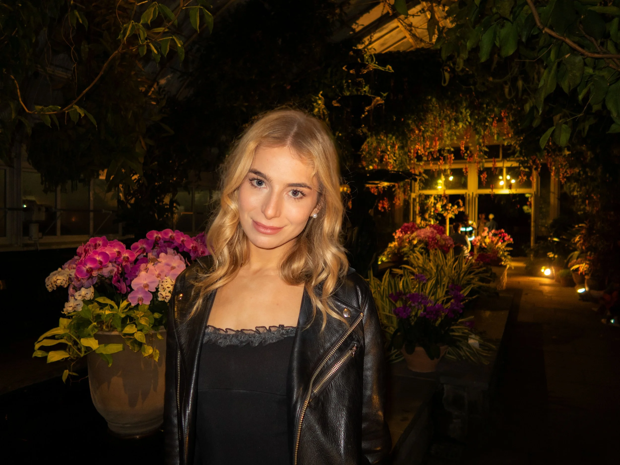 A young woman with blonde hair and a leather jacket standing in a greenhouse or botanical garden filled with flowers and plants at night.