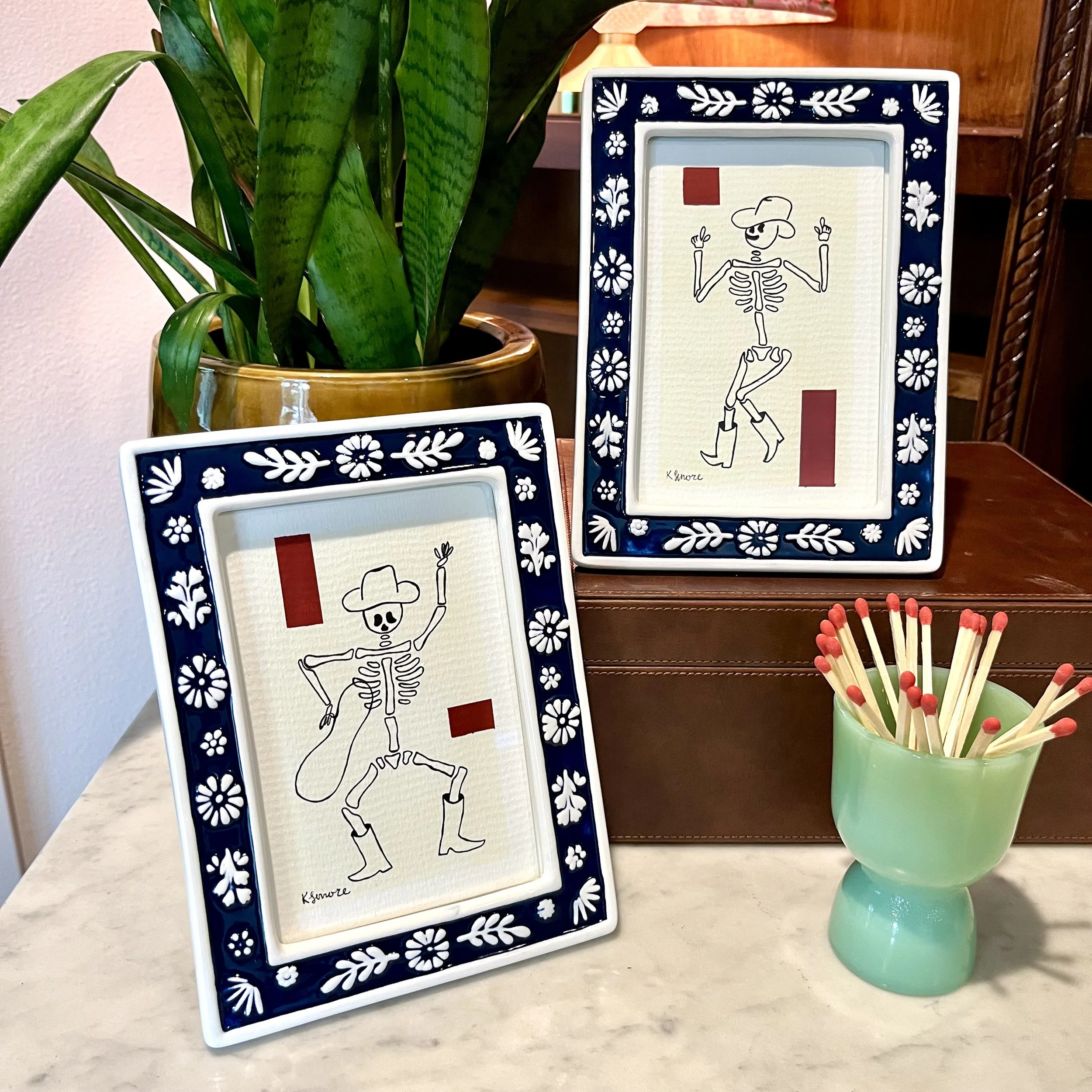 Colorful framed artwork displaying a skeleton in a cowboy hat with raised arms, set against a yellow background with red and blue accents, next to a green vase with a plant and matches in a container.