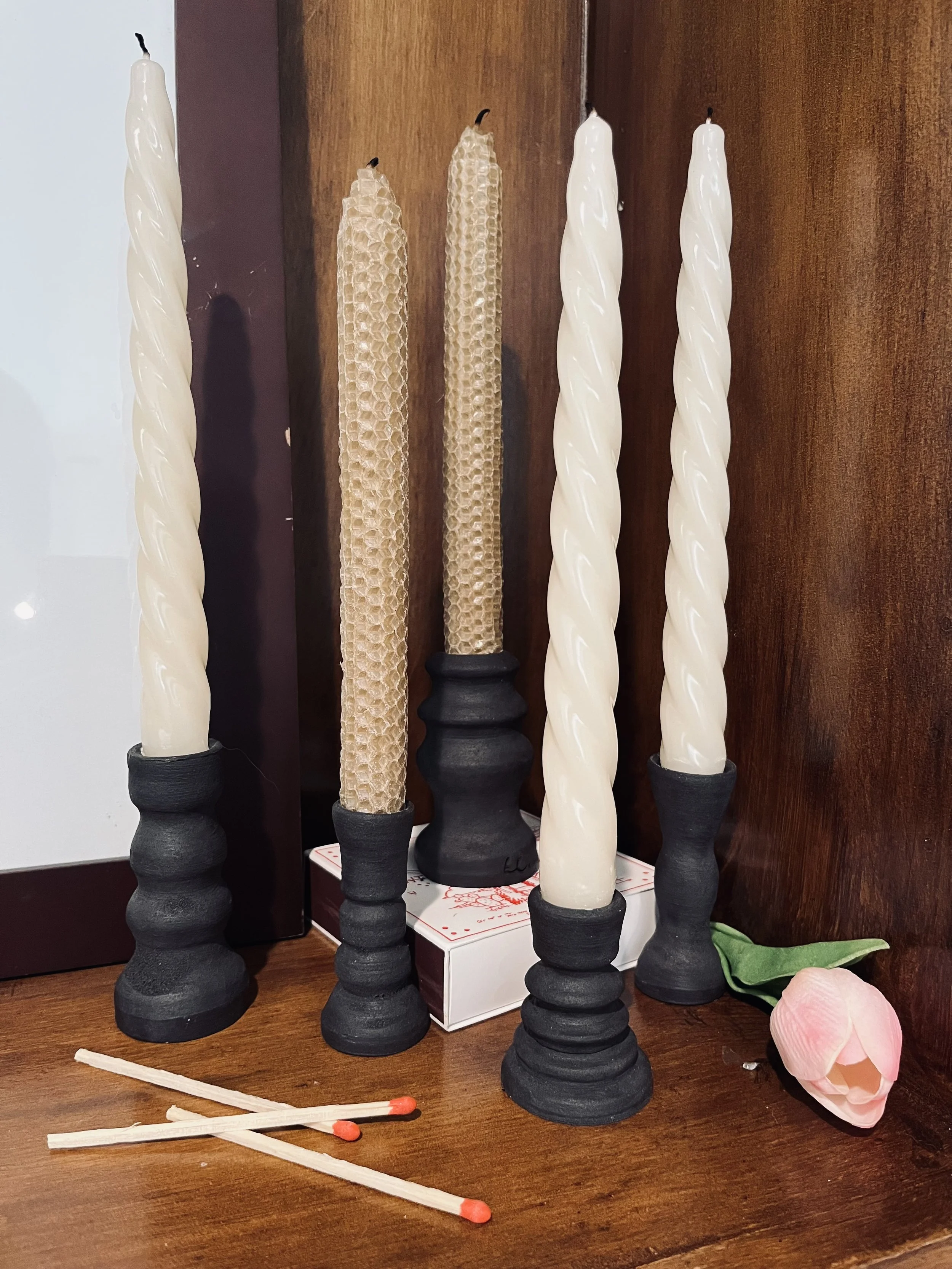 A collection of candles, some with twisted designs and some with honeycomb patterns, displayed on a wooden surface with a pink artificial flower, matchsticks, and a box in the background.