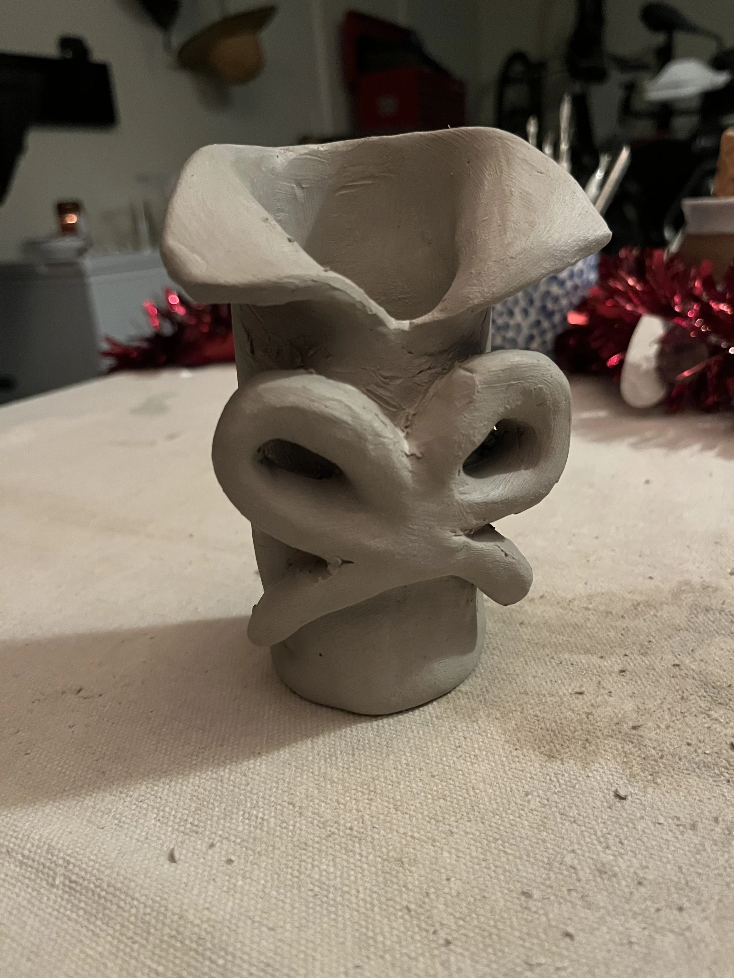 Unfinished ceramic sculpture of a pig's head with exaggerated nostrils, sitting on a beige tablecloth, with Holiday decorations and household items visible in the background.