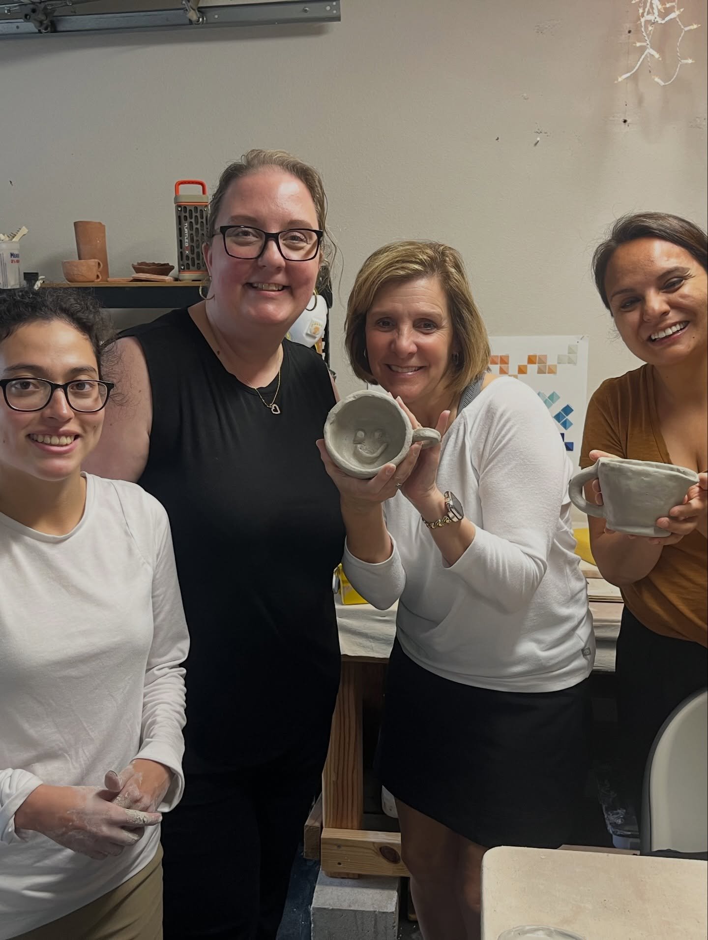 Five women smiling and holding pottery mugs in a workshop. One woman is holding a mug with a smiley face design.