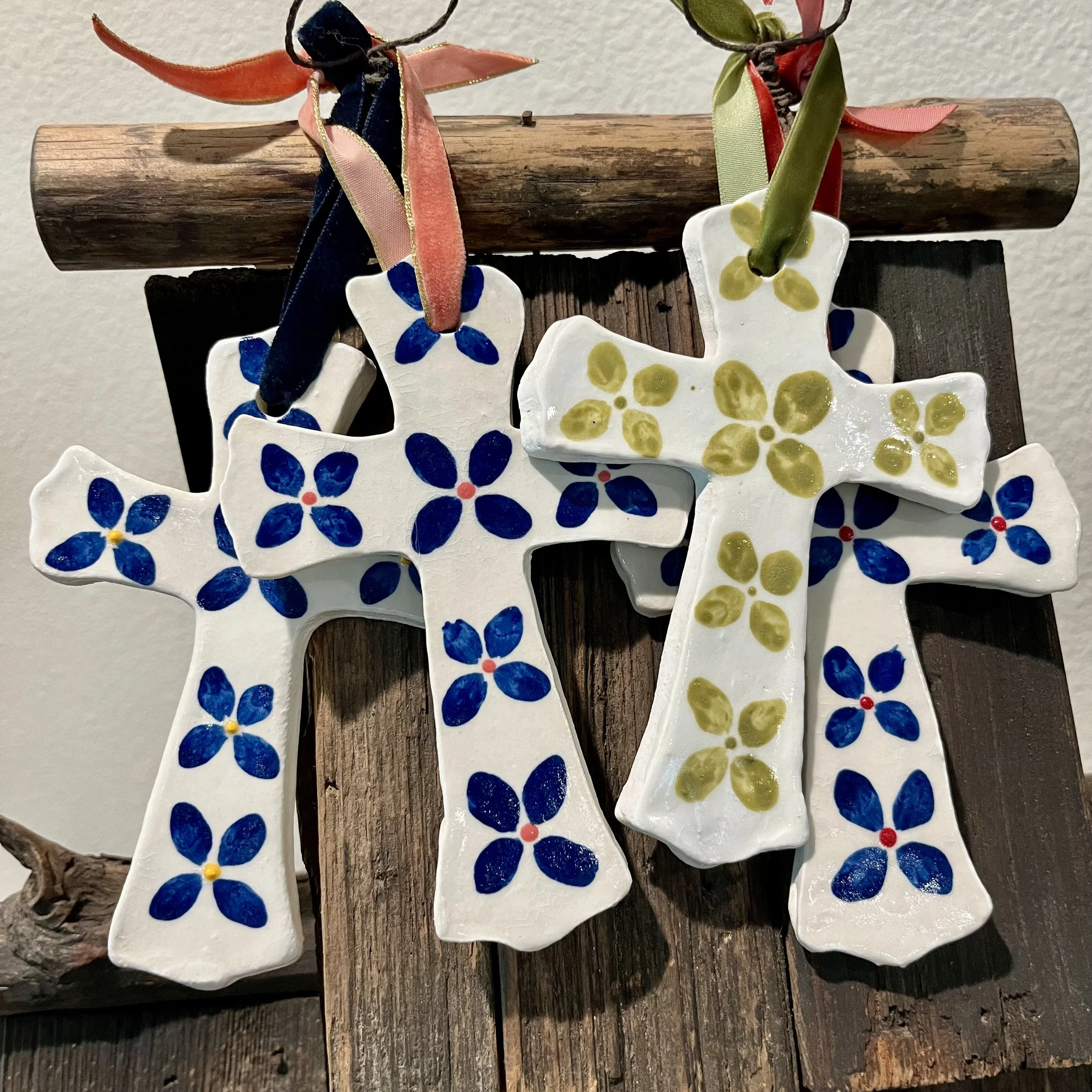 Multiple ceramic snowflake Christmas ornaments with blue and yellow floral patterns hanging from a wooden log, decorated with ribbons, against a plain wall.