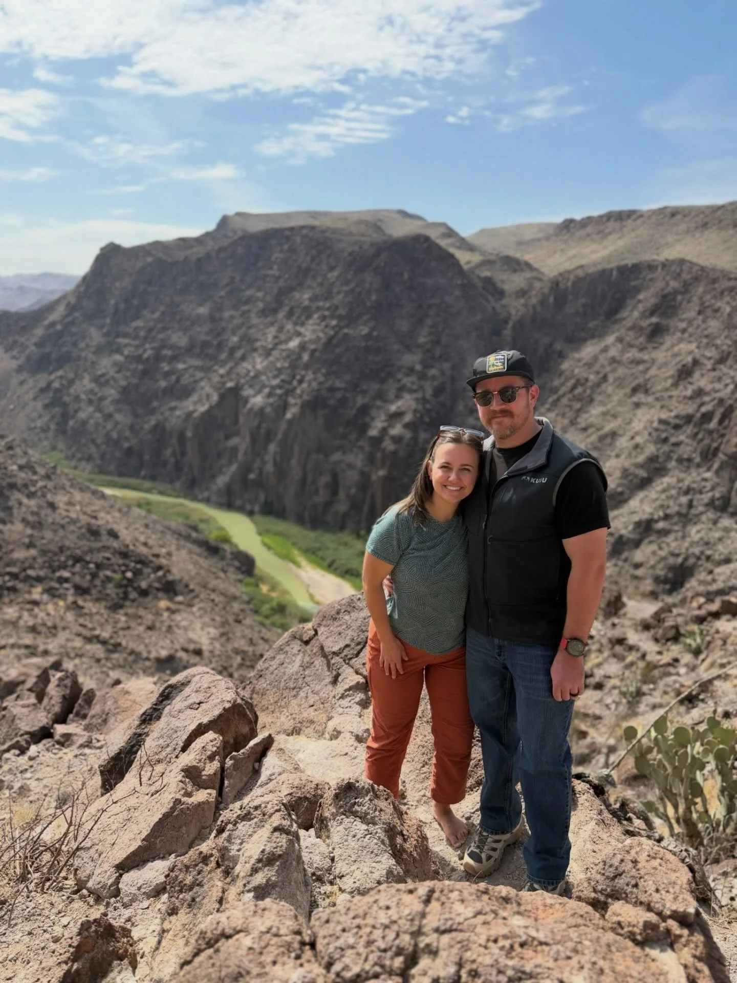 A smiling woman and a man standing close together on rocky terrain in a desert-like landscape with large mountains and a partly cloudy sky in the background.