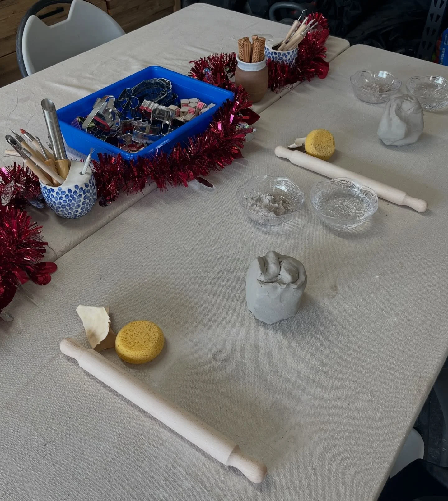 A table decorated with a red tinsel garland, with various glasses, sponges, a rolling pin, baskets of utensils, and small decorative items on it.
