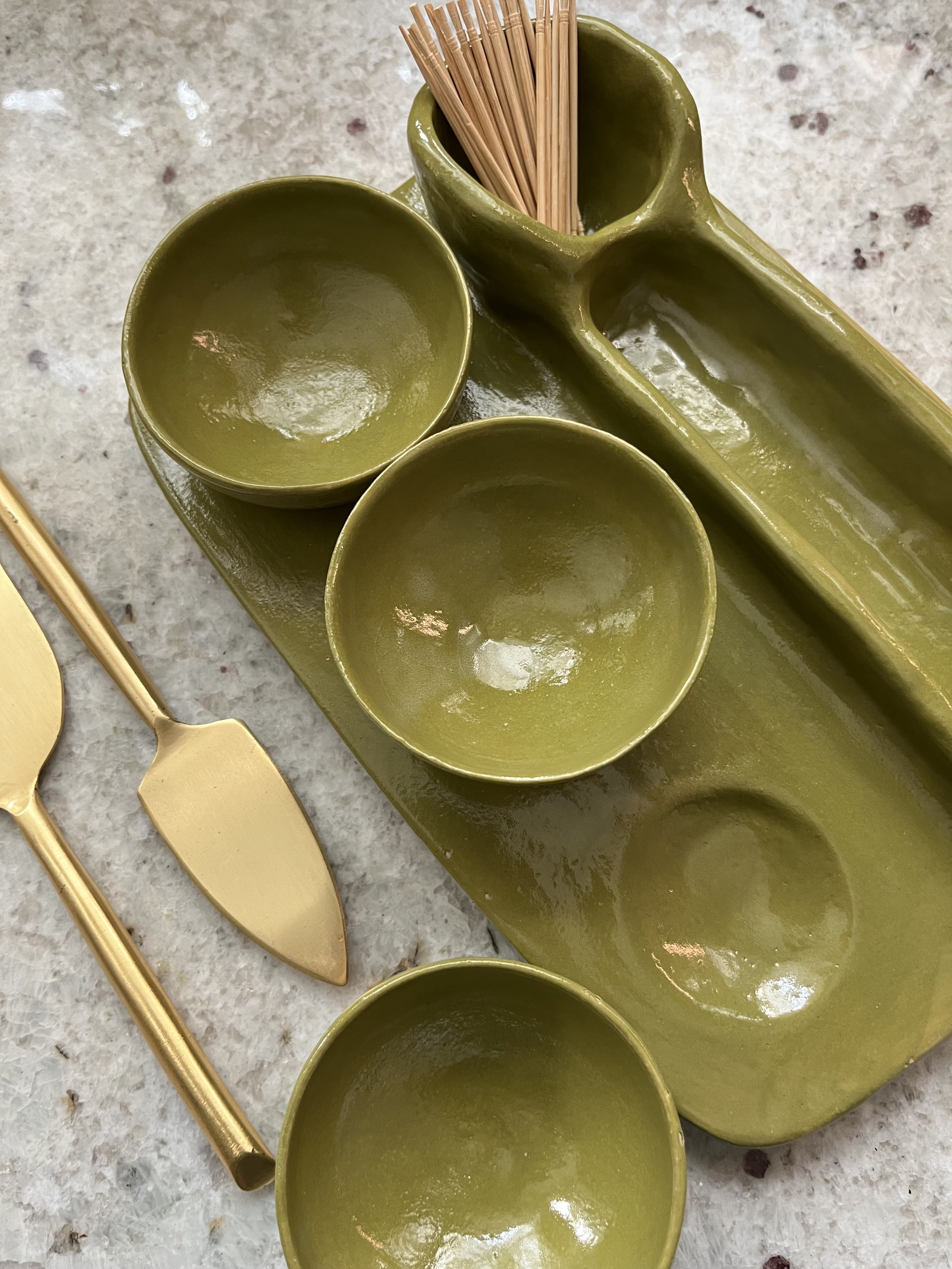 Green ceramic tray holding two green bowls, a cup filled with wooden toothpicks, and a pair of gold-colored scissors, all placed on a gray speckled surface.