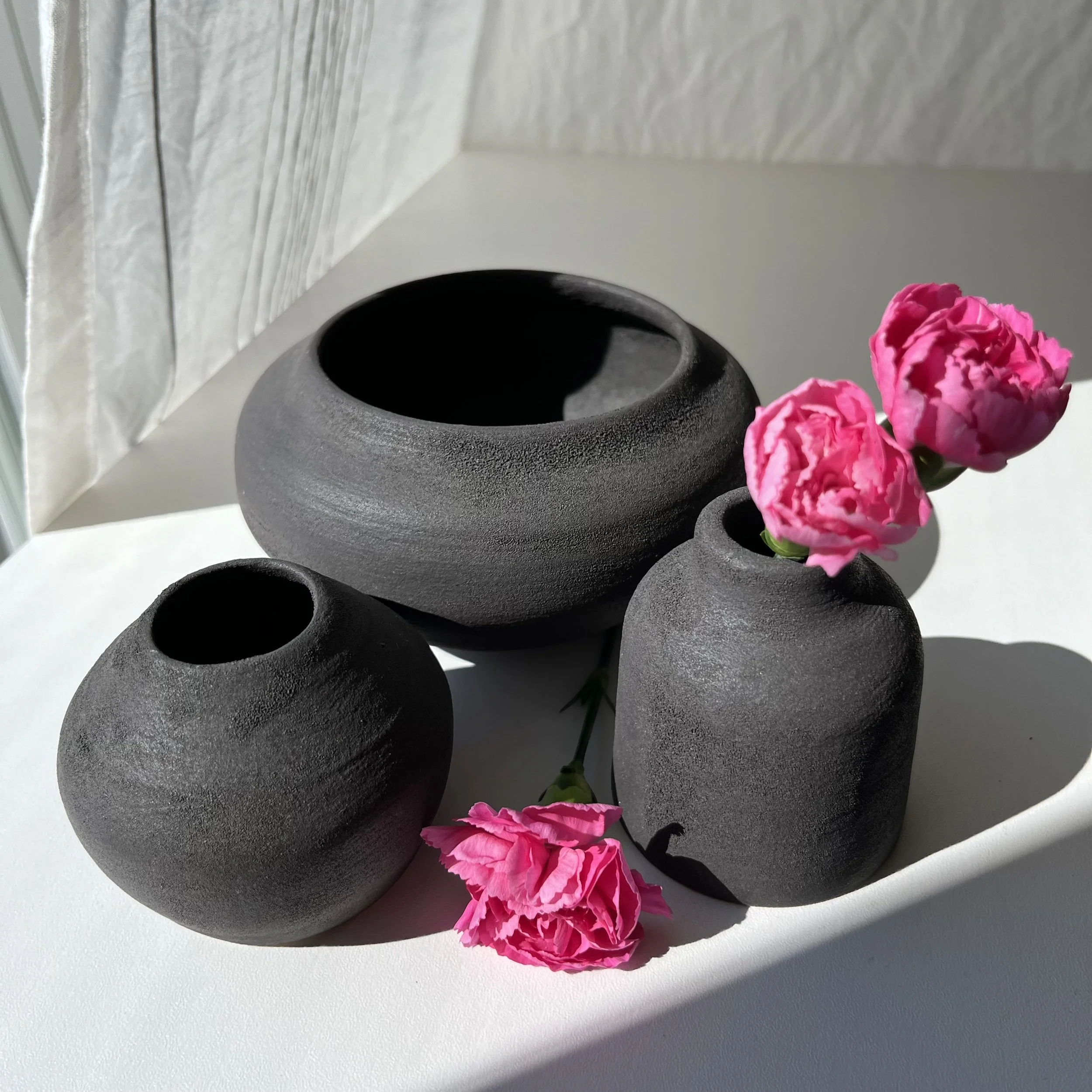 Close-up of a black round vase holding pink carnations, placed on a white surface next to a white wall with a curtain.