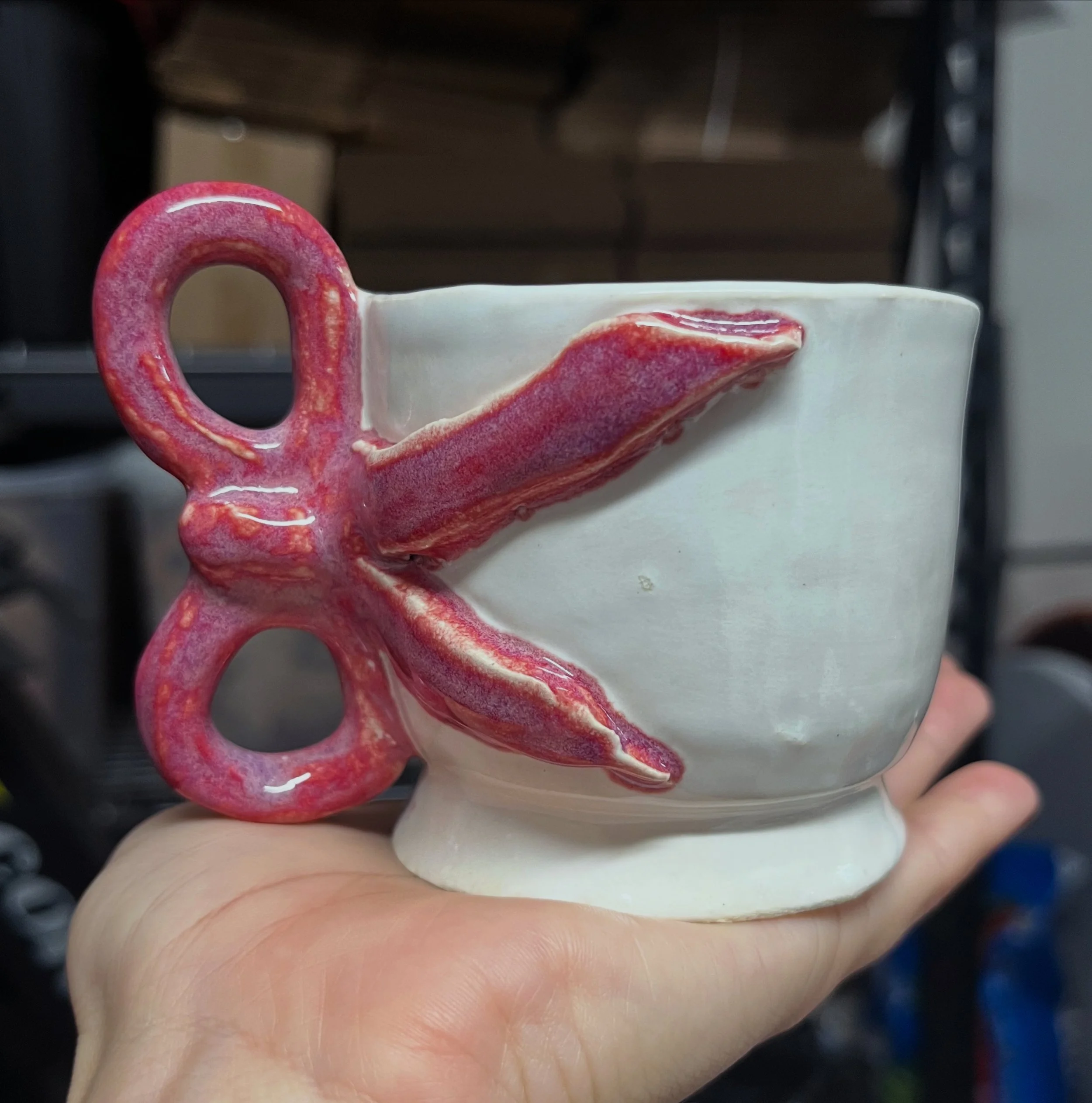 Close-up of a white ceramic mug with a multi-colored, textured pink handle and decorative pink and red accents around the rim, held by a person with a ring on their finger.