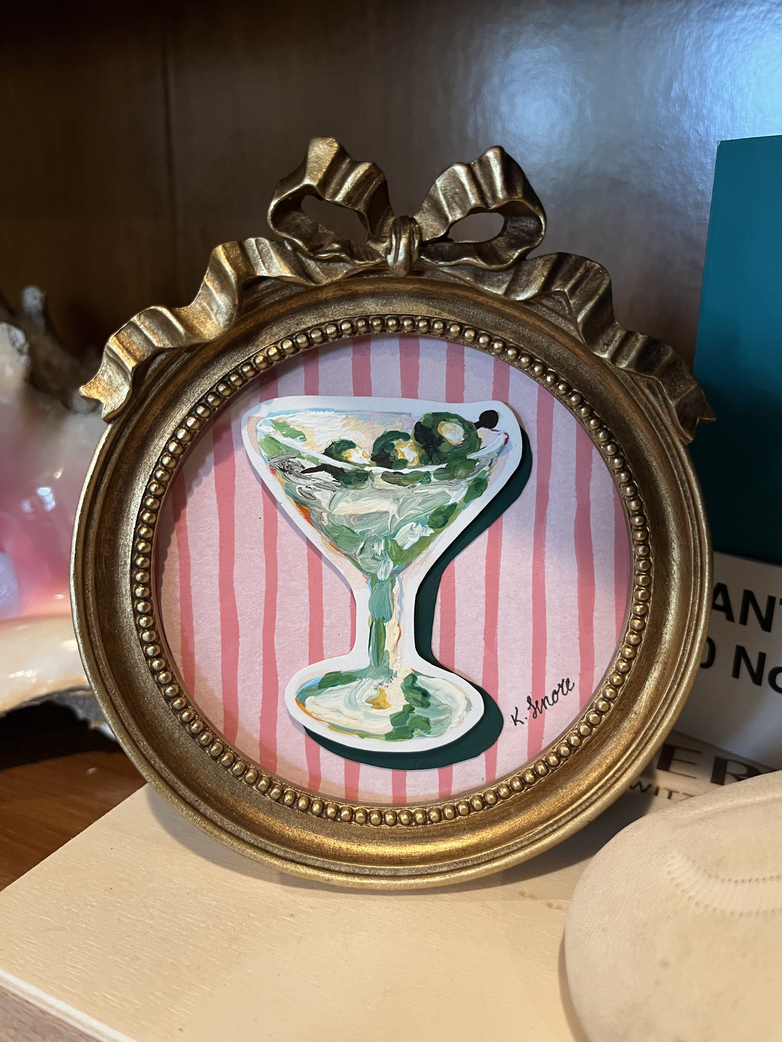 A gold ornate picture frame with a pink striped background. Inside the frame is a painted illustration of a martini glass with green olives, signed 'K. Smart' in the lower right corner.