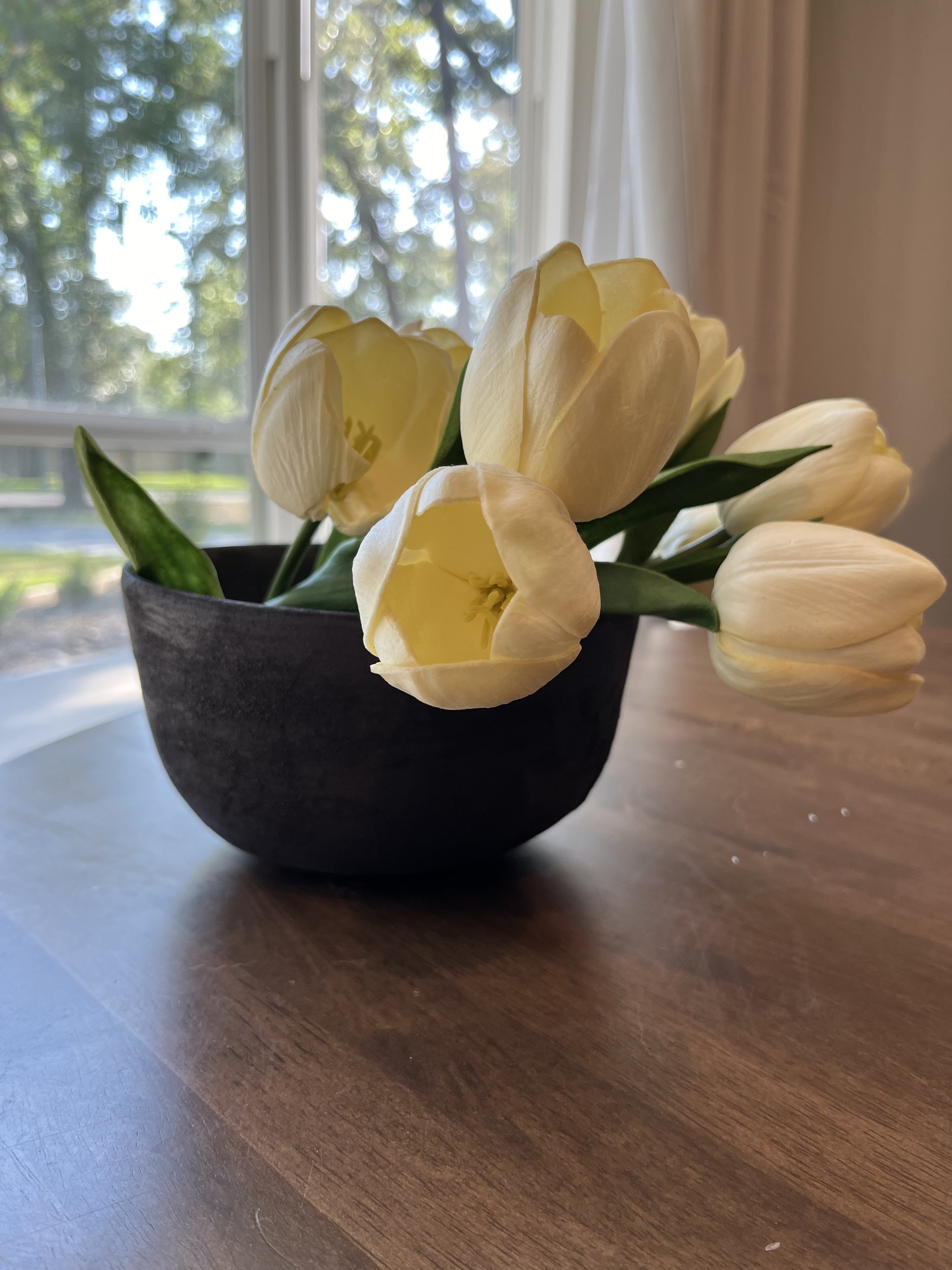 Yellow tulips in a black pot on a wooden table with a window showing trees in the background.