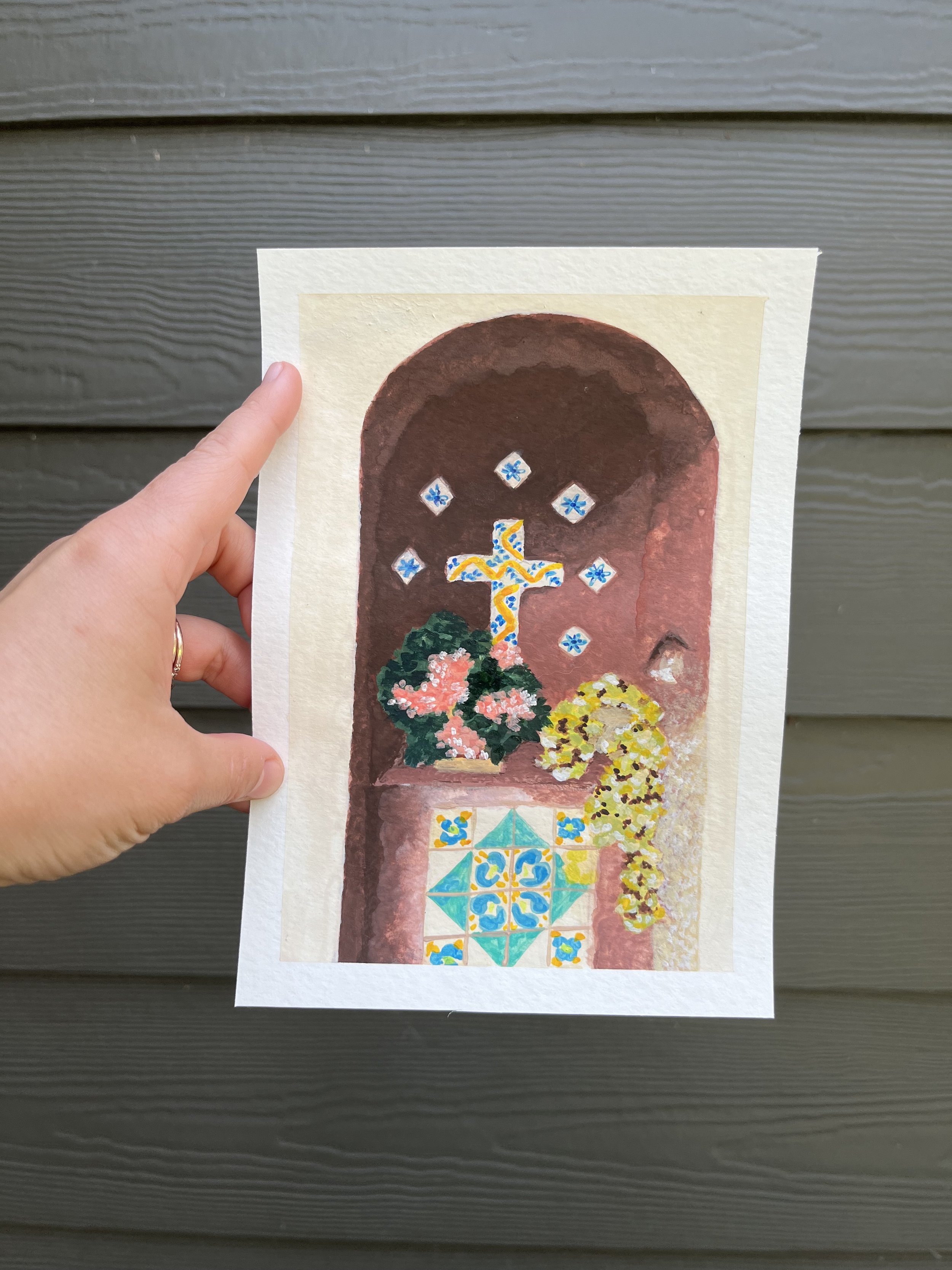 A hand holding a piece of artwork featuring a religious or decorative cross, surrounded by colorful flowers and a patterned tile design at the bottom, against a background of a gray, horizontal wooden surface.
