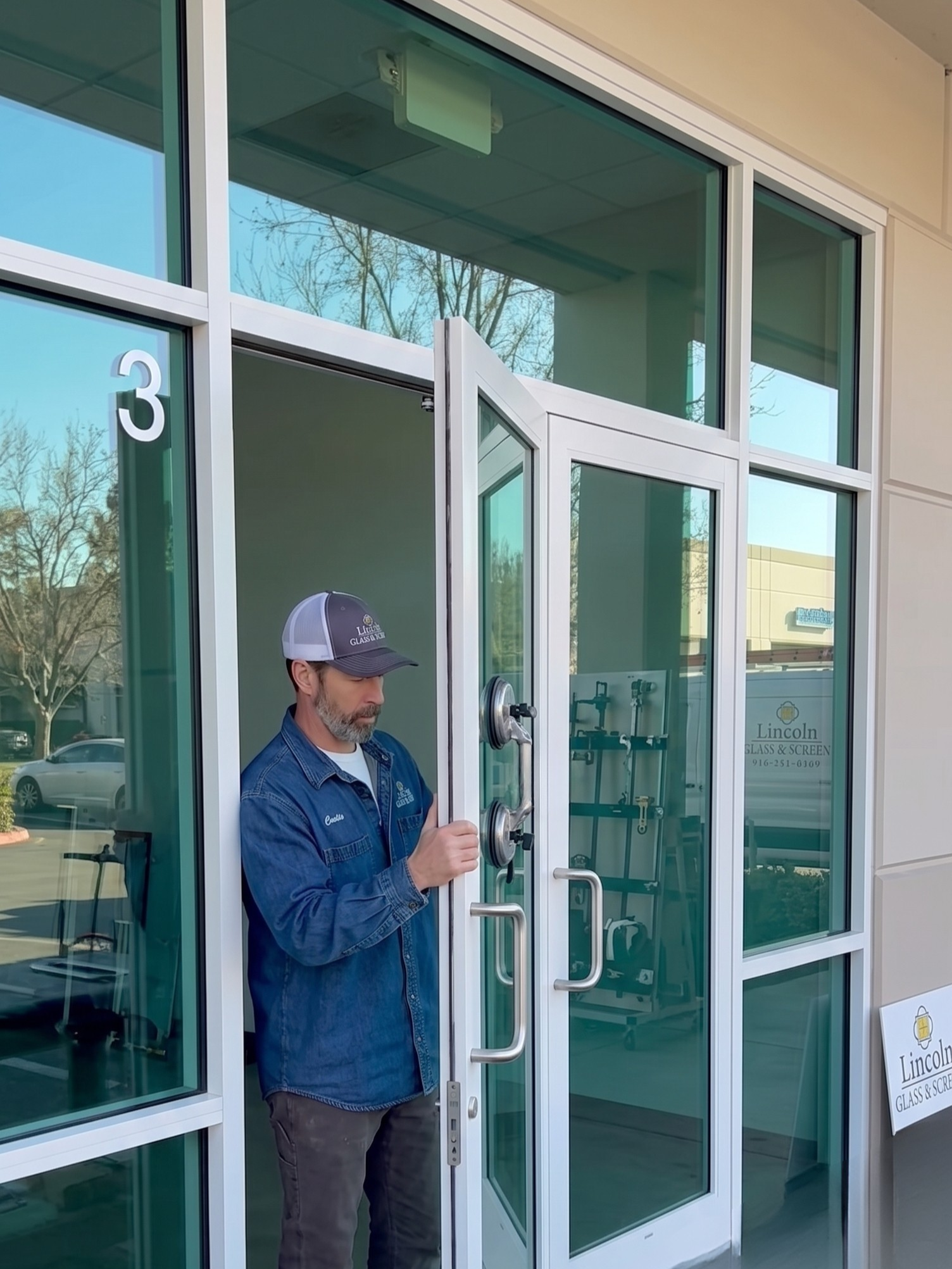 Man in blue jacket and grey cap cleaning glass door with a handheld squeegee. The door has a sign that reads 'Lincoln Glass & Screen'.