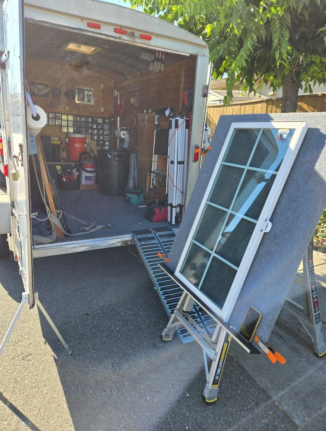 Photo of a trailer with tools, a window panel, and other equipment inside and outside, set up for work or transport.