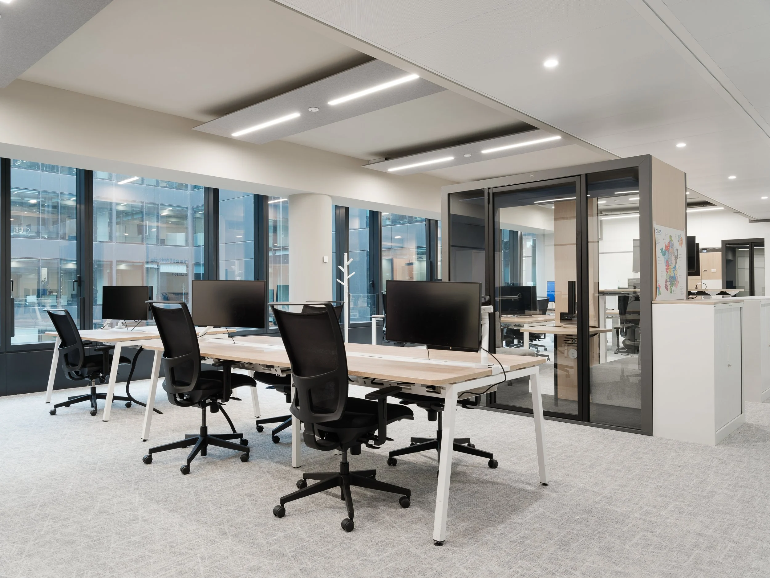 Un espace de bureau moderne avec plusieurs tables, chaises, écrans d'ordinateur, des grandes fenêtres laissant entrer la lumière naturelle, des murs blancs et un plafond lumineux.