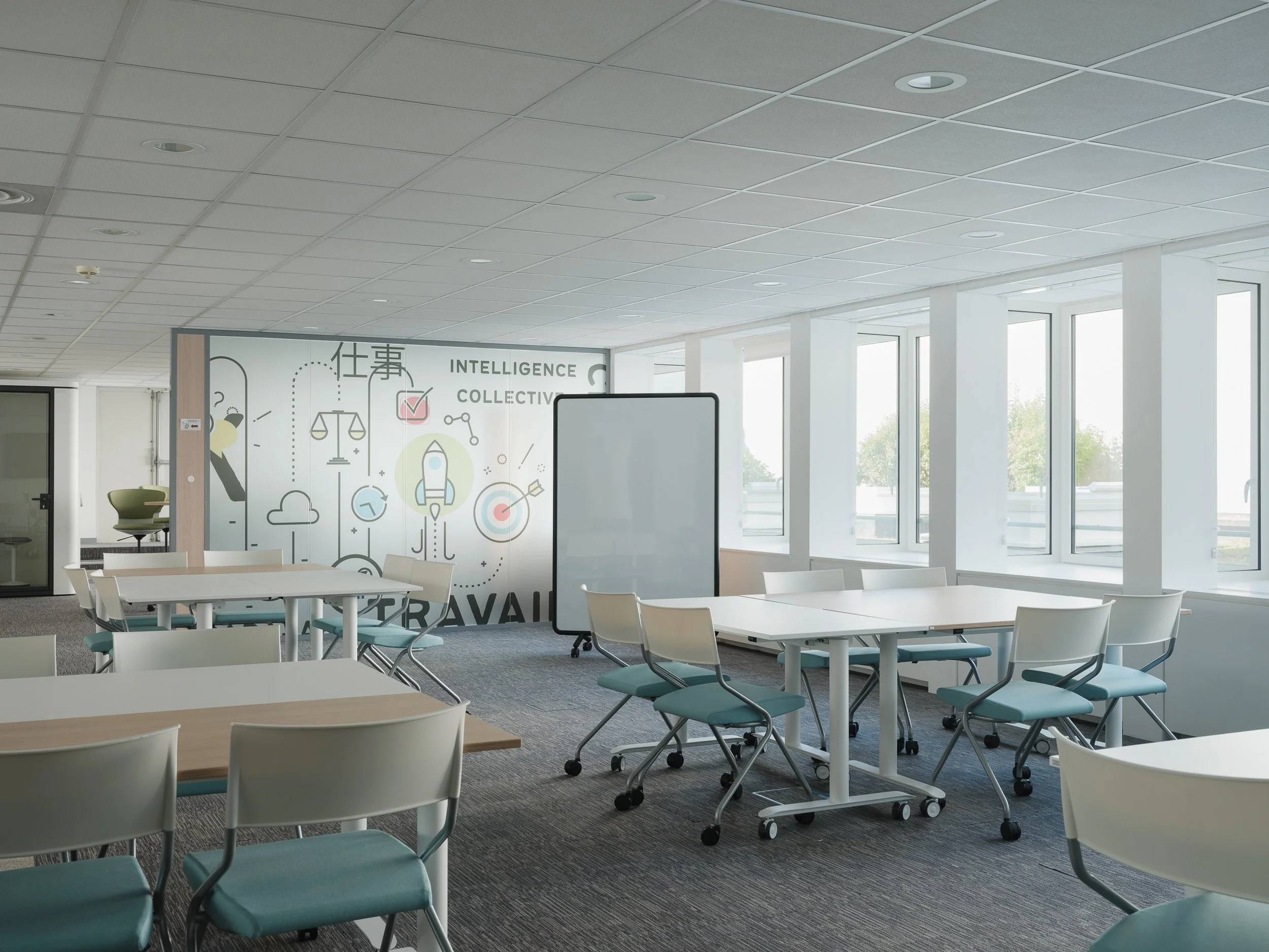 Salle de réunion ou espace de coworking avec plusieurs tables et chaises, grands fenêtres laissant entrer la lumière naturelle, mur avec dessin sur le thème de l'intelligence collective, ciel dégagé visible à l'extérieur.