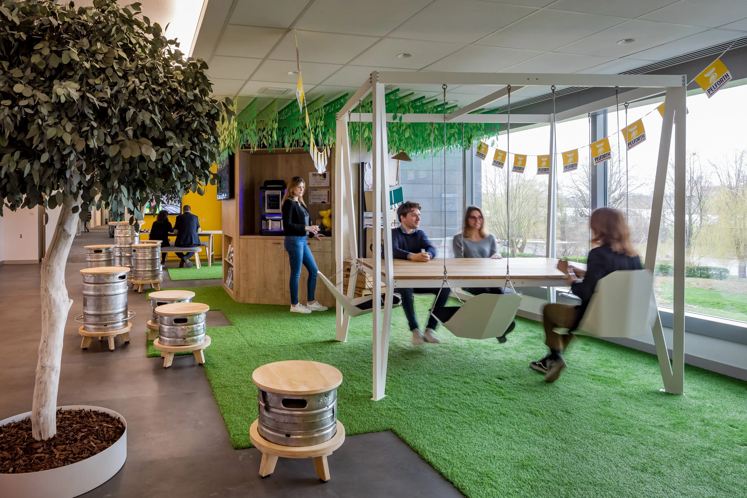 Espace de coworking avec des plantes, des tables, des cafés en métal, des sièges confortables et une grande baie vitrée laissant entrer la lumière naturelle.
