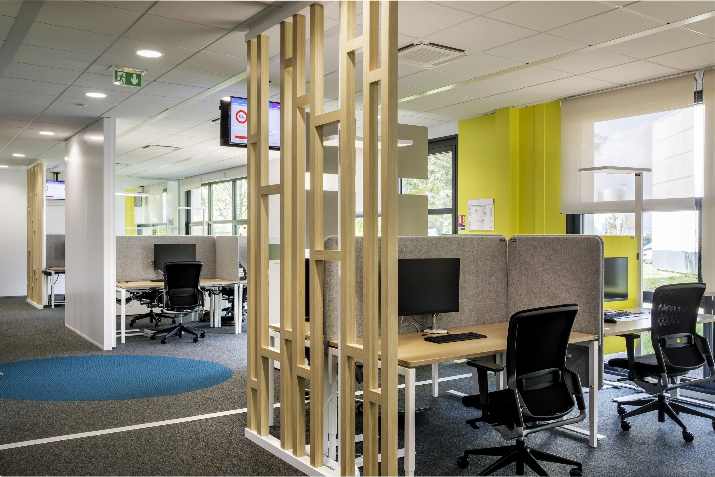 Espace de bureau ouvert avec des stations de travail séparées par des parois en tissu gris, une cloison en bois clair, des fenêtres laissant entrer la lumière naturelle, et un plafond blanc avec des lumières intégrées.