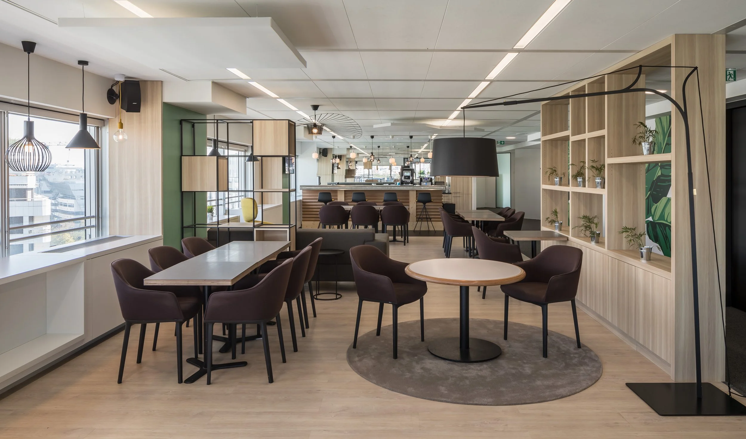 Intérieur d'un espace de coworking ou café avec plusieurs tables et chaises, étagère décorative avec plantes, éclairage suspendu, grande fenêtre avec vue sur la ville.