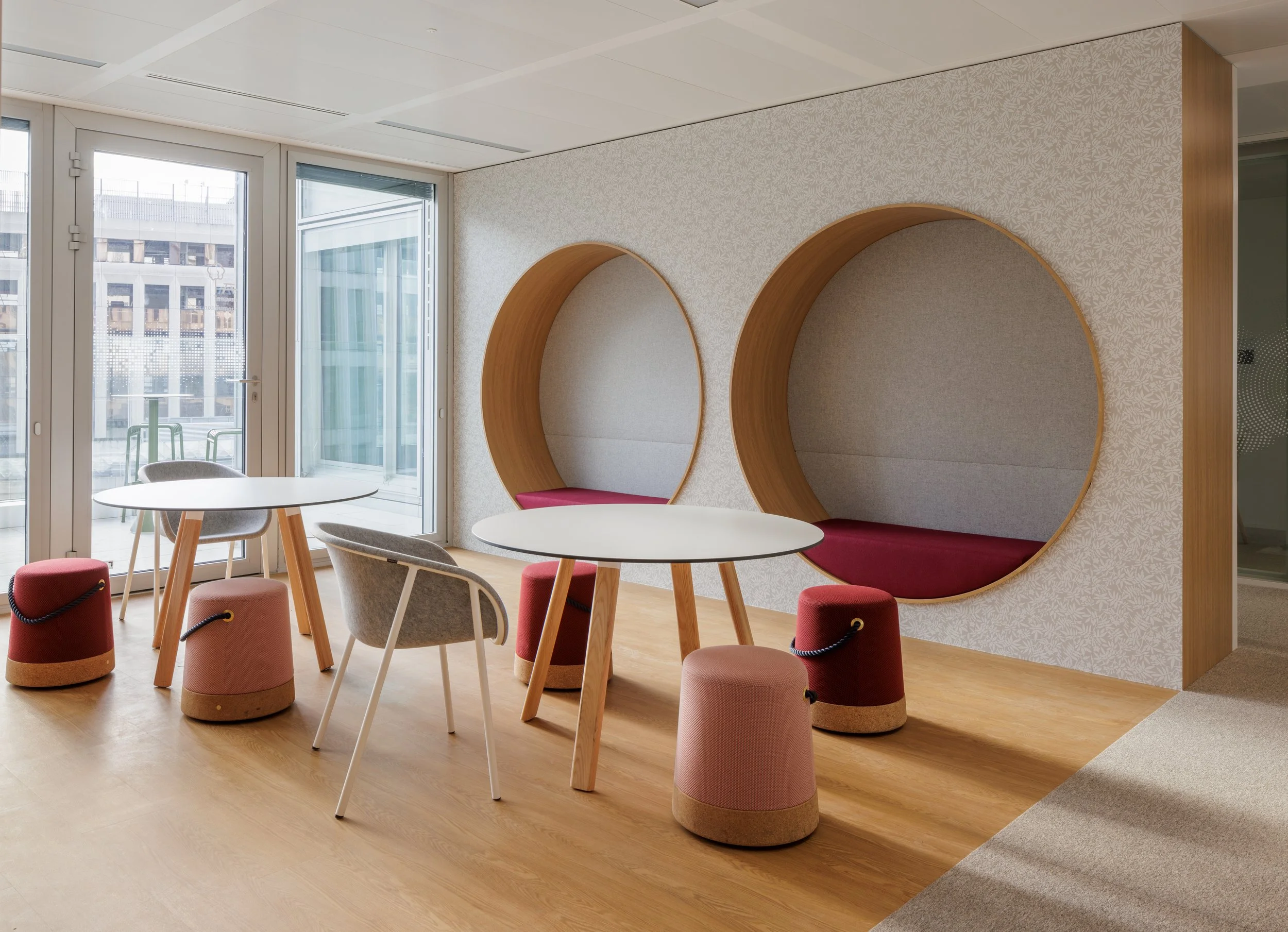 Intérieur moderne d'une salle de réunion avec des tables rondes, des fauteuils et des poufs, un mur décoratif avec deux formes circulaires en bois, grande fenestration laissant entrer la lumière naturelle.