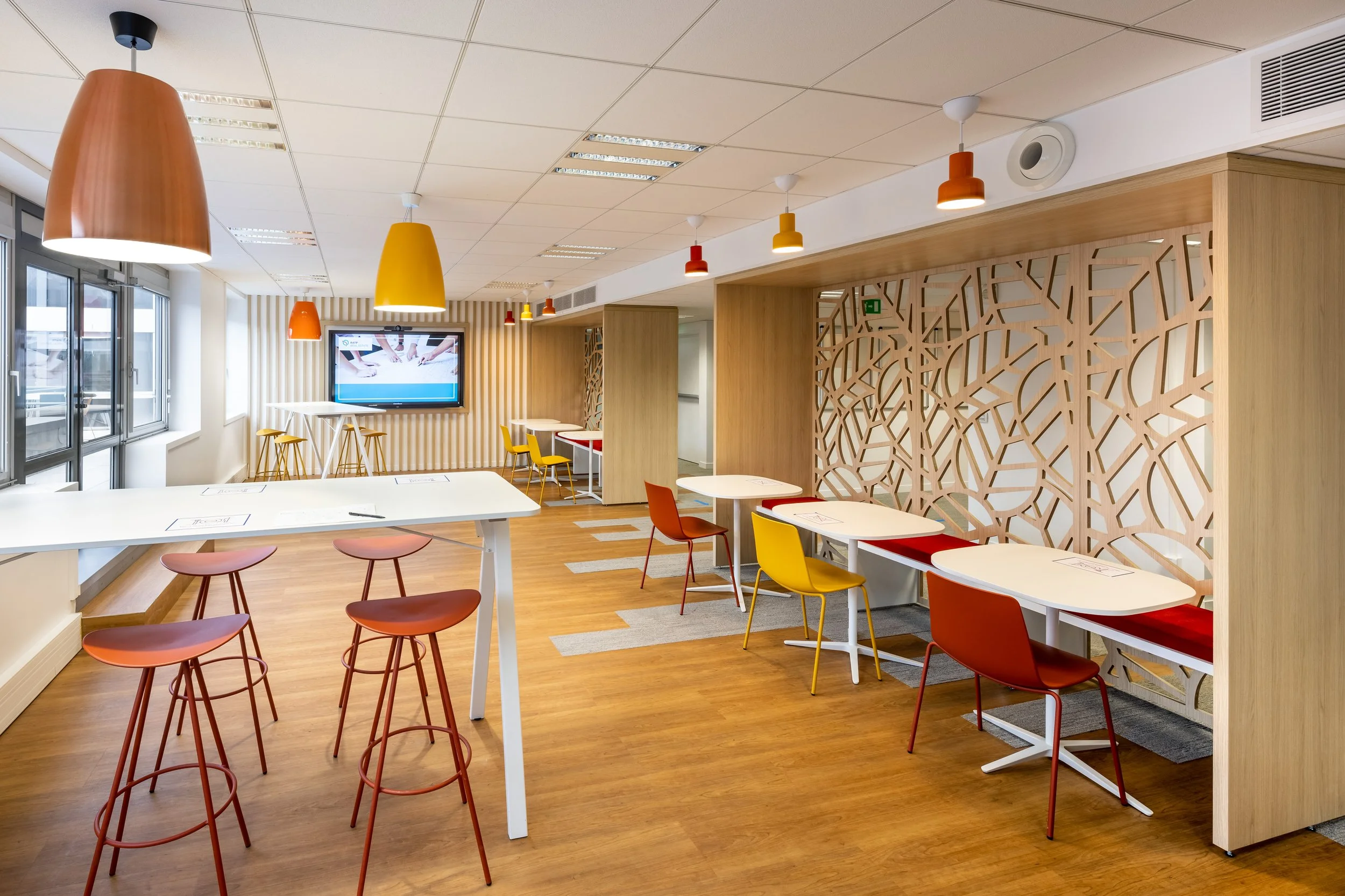 Un espace de bureau moderne avec des tables blanches, des chaises jaunes, oranges et rouges, des lampes suspendues de couleurs différentes, une grande télévision et des murs en bois décoratif avec un motif géométrique. La pièce est lumineuse grâce aux fenêtres proches.