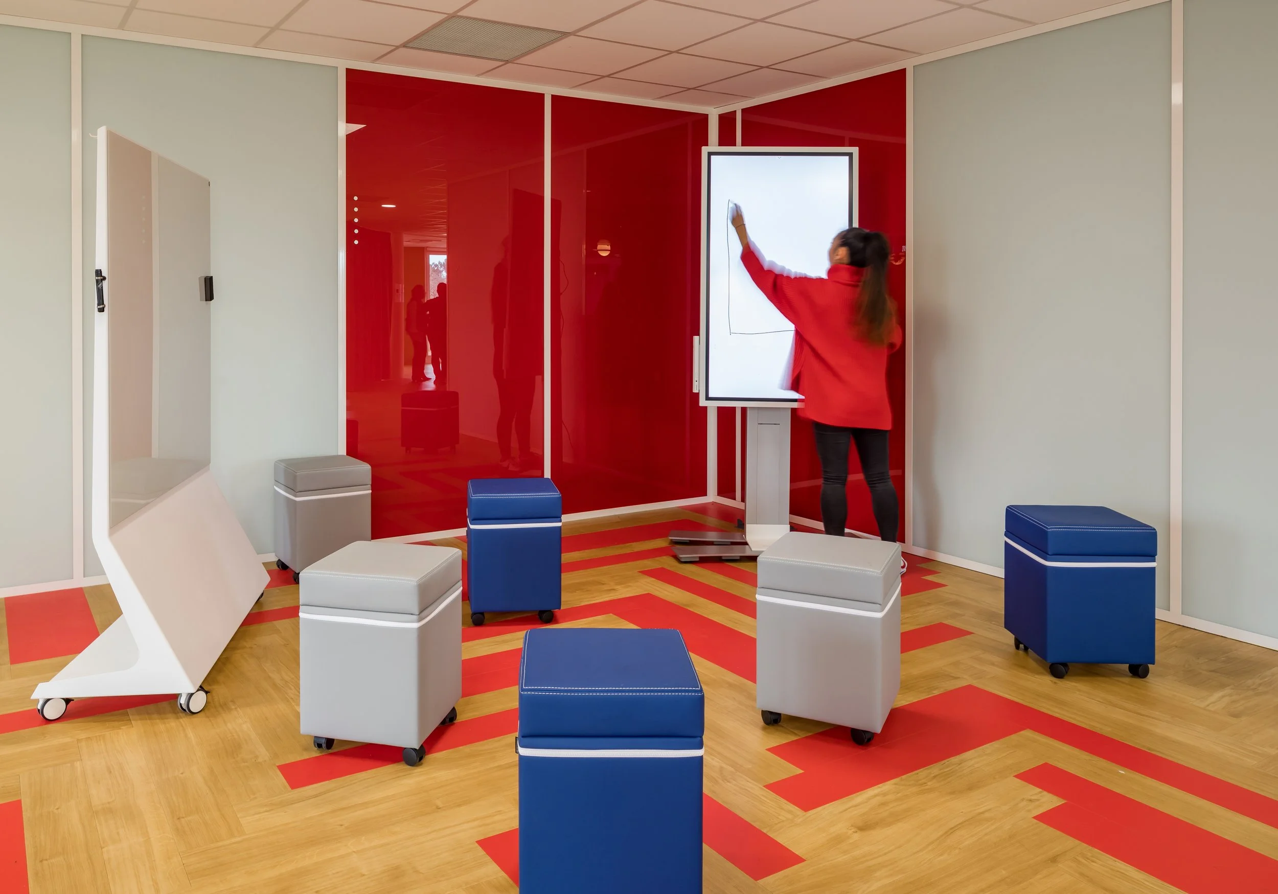 Une femme en rouge écrit sur un tableau blanc numérique dans une salle moderne avec des mobiliers carrés bleus et gris sur un sol en bois, derrière une paroi rouge vernie.