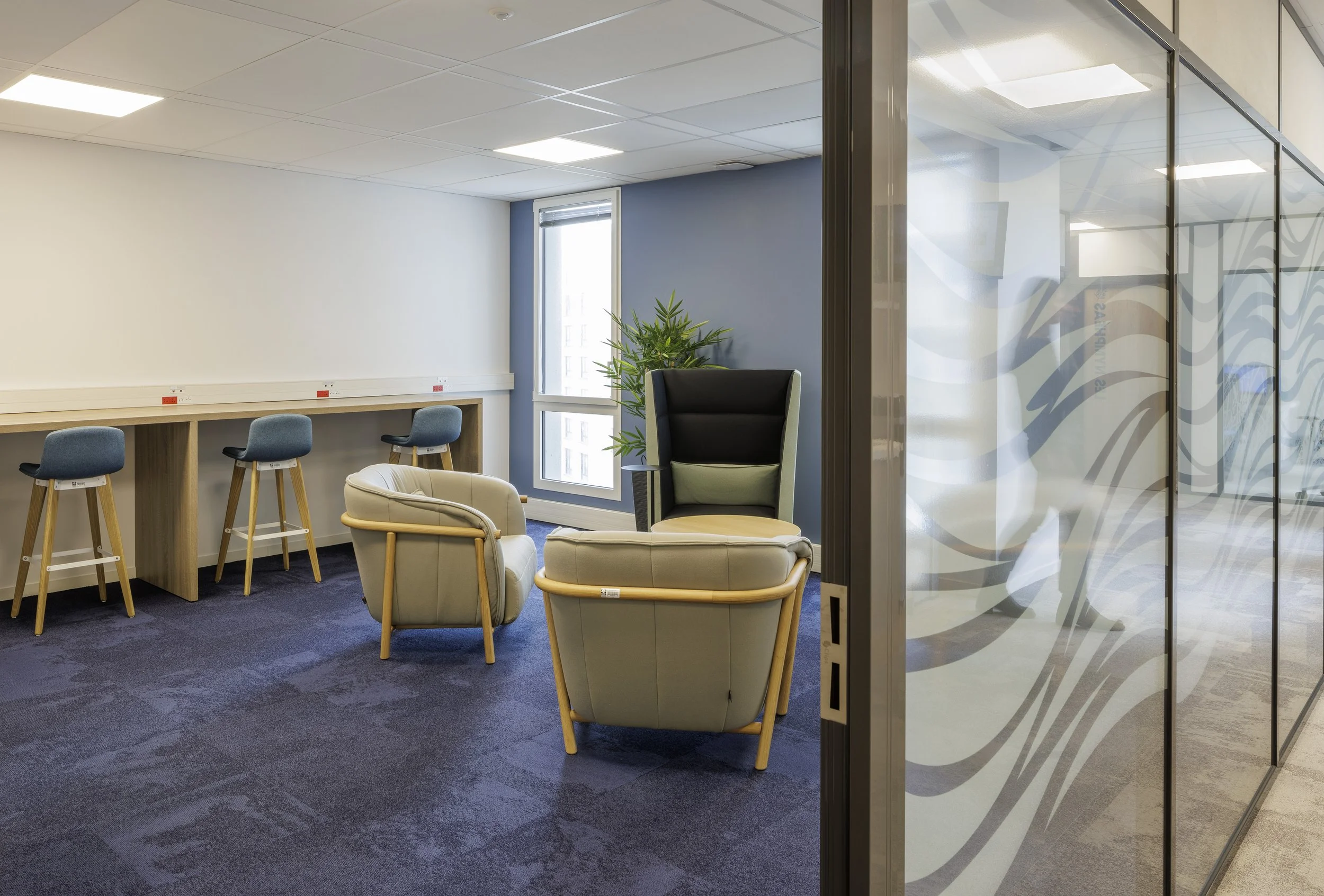 Une salle de réunion ou espace de travail avec des fauteuils, une table avec des barres de câbles électriques, une plante verte, et une fenêtre. La pièce est séparée d'un espace voisin par une cloison en verre avec un motif décoratif.