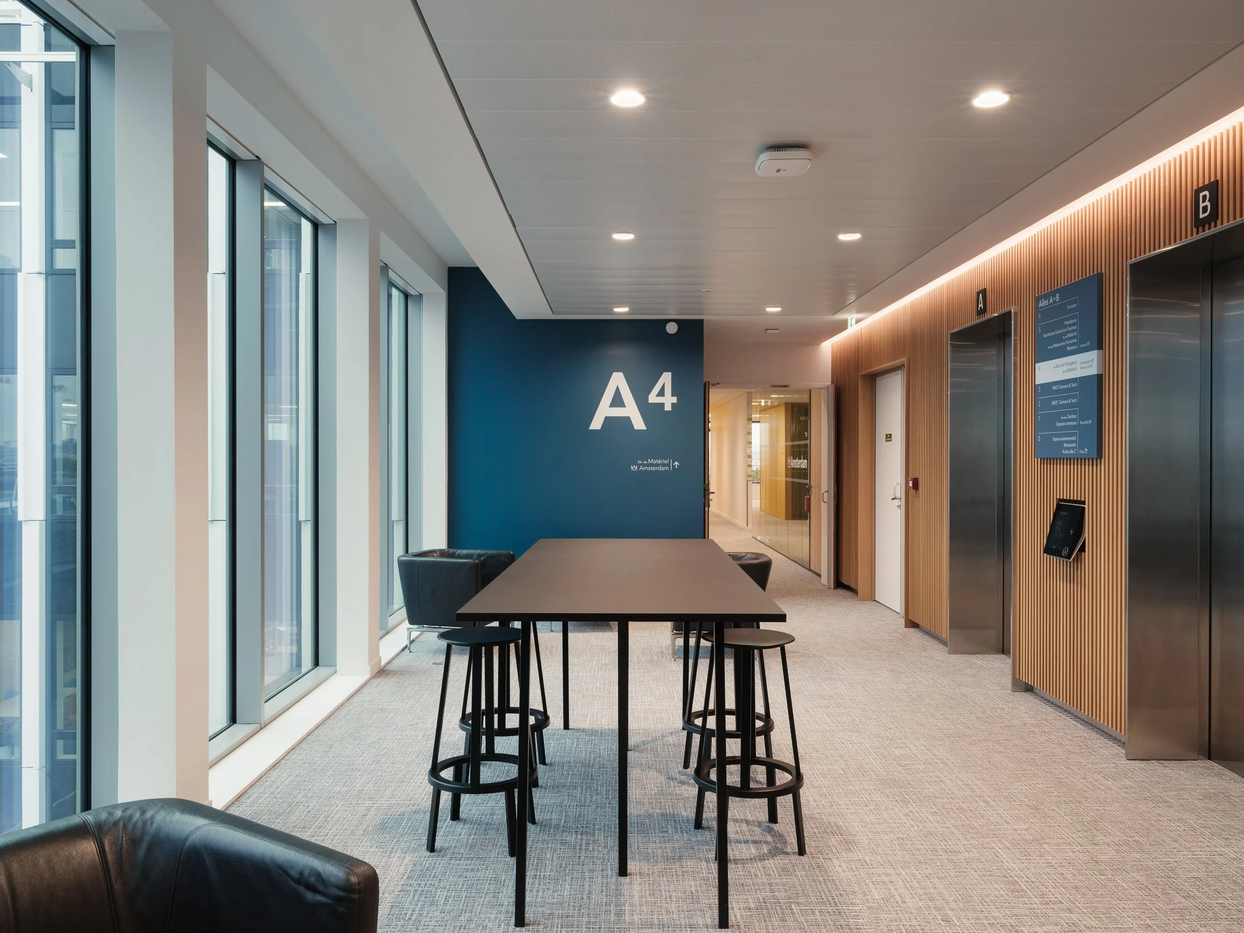 Salle d'attente moderne dans un bâtiment, avec un mur bleu portant le signe A4, des fenêtres côté gauche laissant entrer la lumière naturelle, une table haute avec quatre tabourets, et des ascenseurs avec un panneau d'information numérique sur le mur en bois.