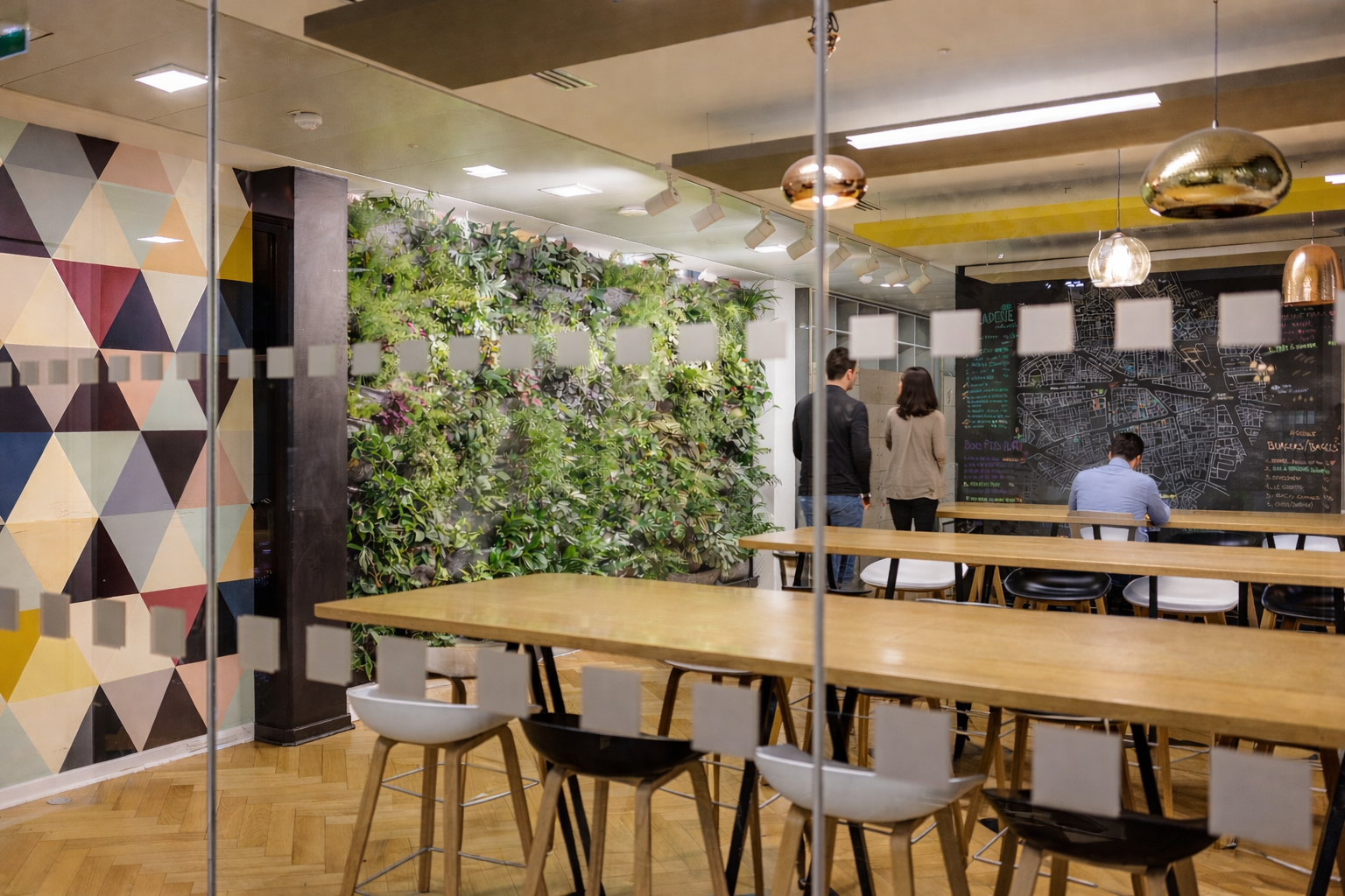 Intérieur d'un café ou d'un restaurant moderne avec des tables en bois, des chaises colorées, un mur végétal et un tableau noir avec une carte. Plusieurs personnes discutent ou travaillent.