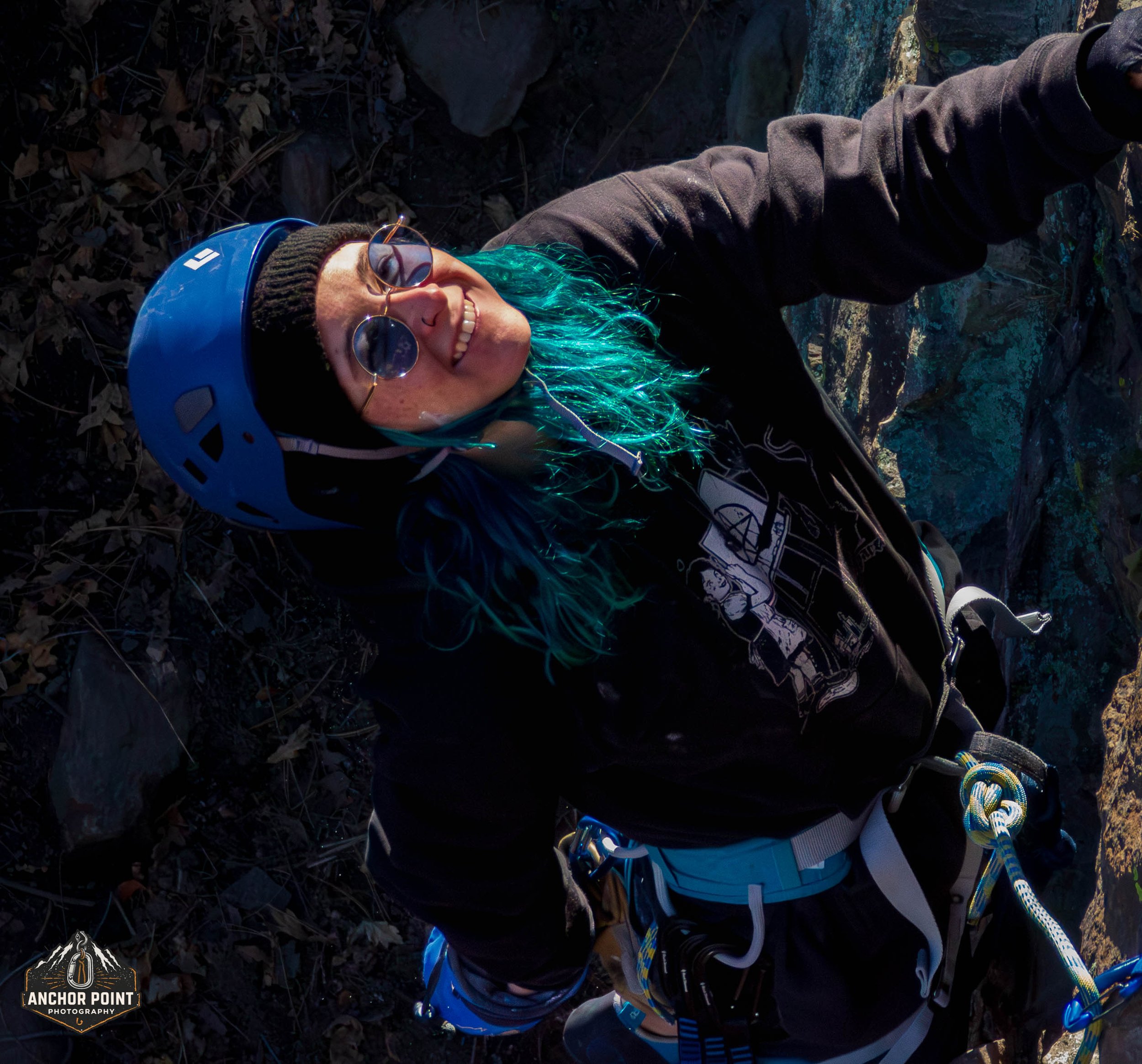 A woman with teal hair, wearing sunglasses, a black beanie, and a blue helmet, rocks climbing outdoors, smiling and looking up.