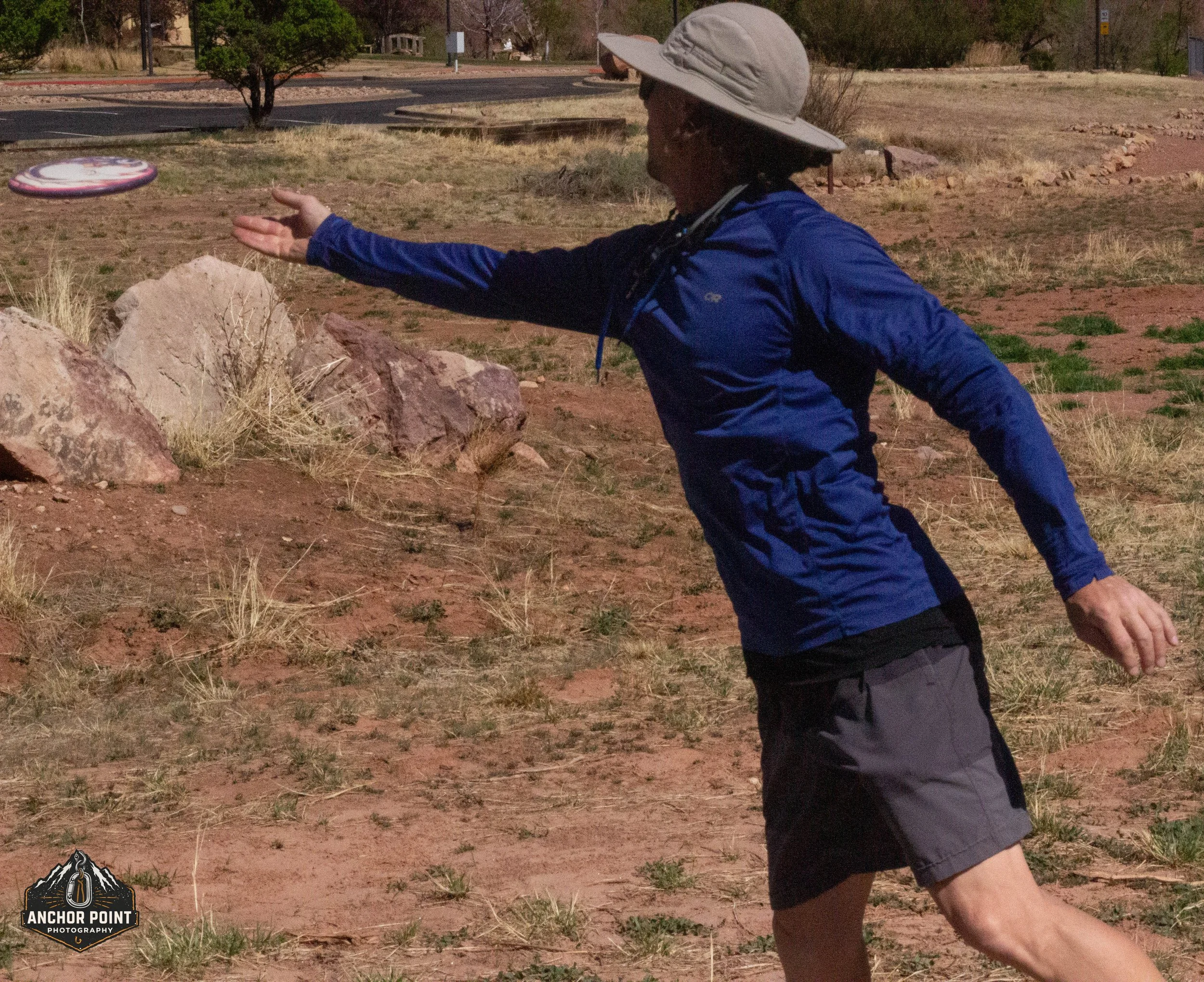 A person wearing a wide-brimmed hat, a blue long-sleeve shirt, and gray shorts is throwing a frisbee in an outdoor, desert-like area with rocks and sparse dry grass.