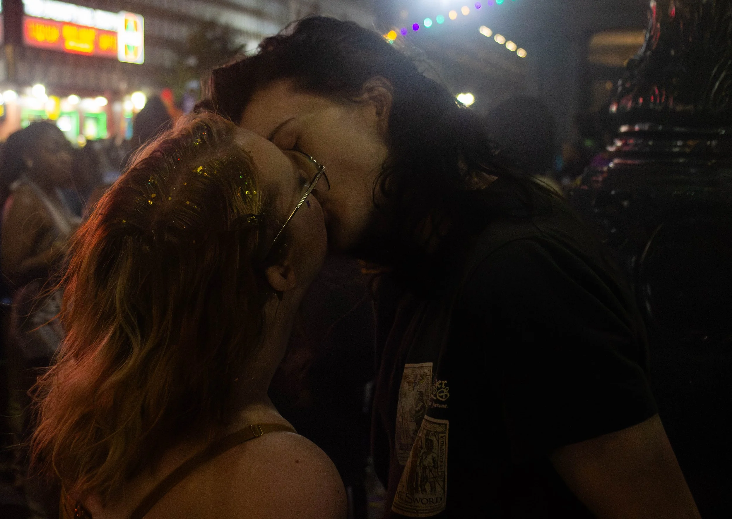 A couple sharing a kiss at a nighttime event or party, with blurred lights and other people in the background.