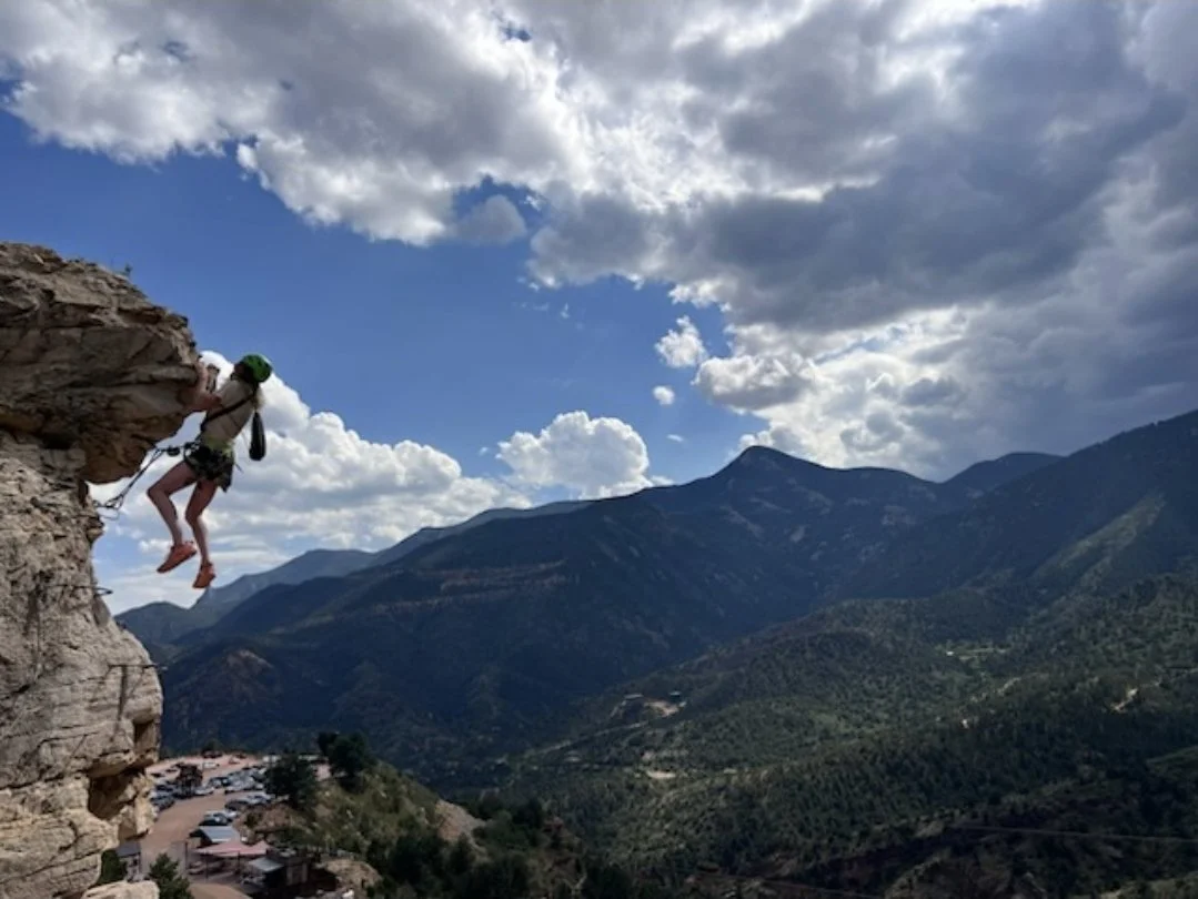 A person rock climbing on a cliff with a mountainous landscape and partly cloudy sky in the background.