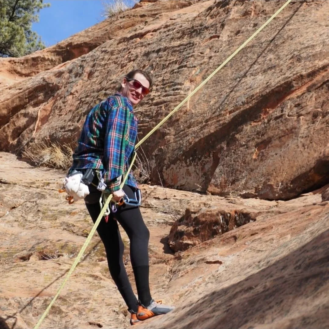 A person wearing a plaid shirt, black leggings, climbing shoes, and red sunglasses is rock climbing on a red sandstone surface. They are secured with a climbing harness and safety ropes, and are smiling at the camera.