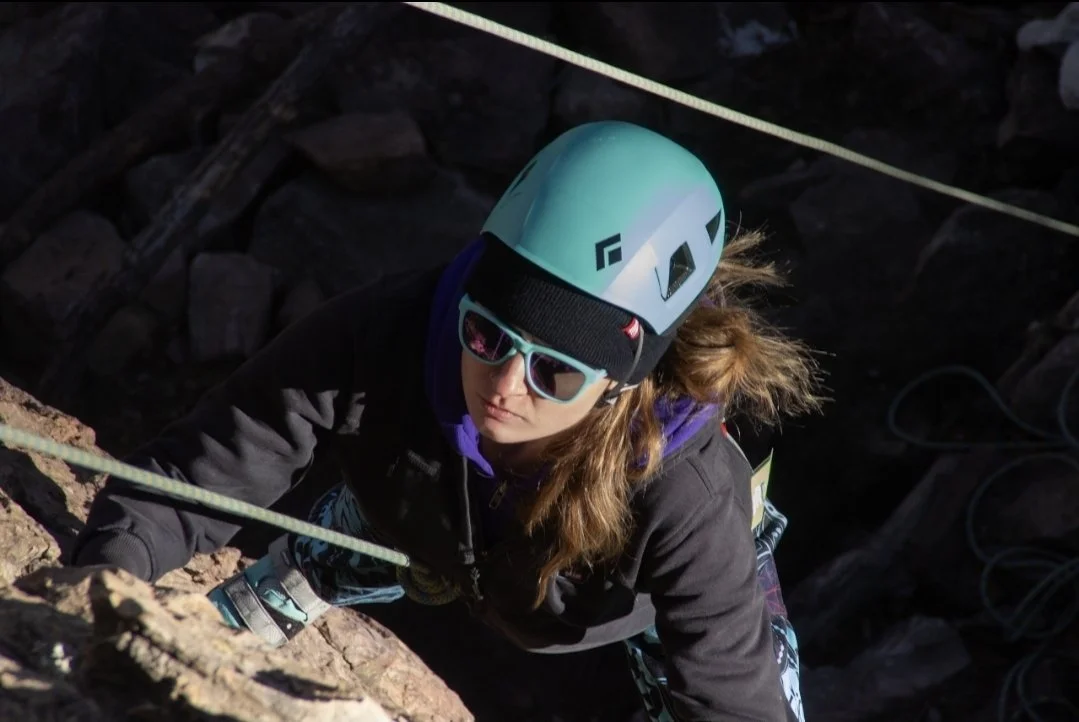 A woman rock climbing outdoors, wearing a blue helmet, sunglasses, and a black jacket.