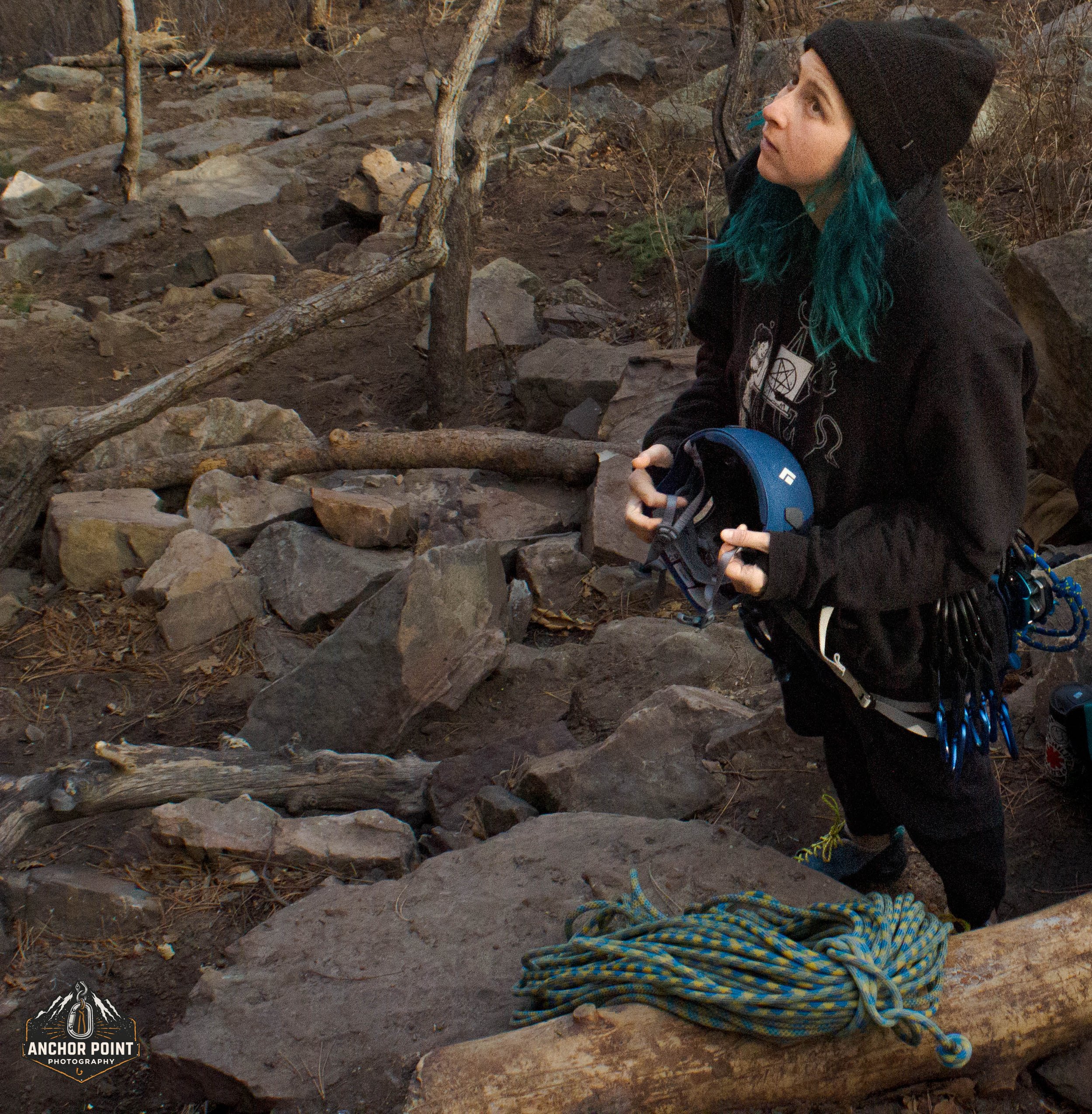 A woman with teal hair wearing a black beanie and black hoodie stands outdoors among rocks and fallen logs, holding a climbing helmet in her hands. There is climbing gear hanging from her belt. A rolled-up blue and yellow rope is on a log in front of her. The background shows a rocky, wooded area.