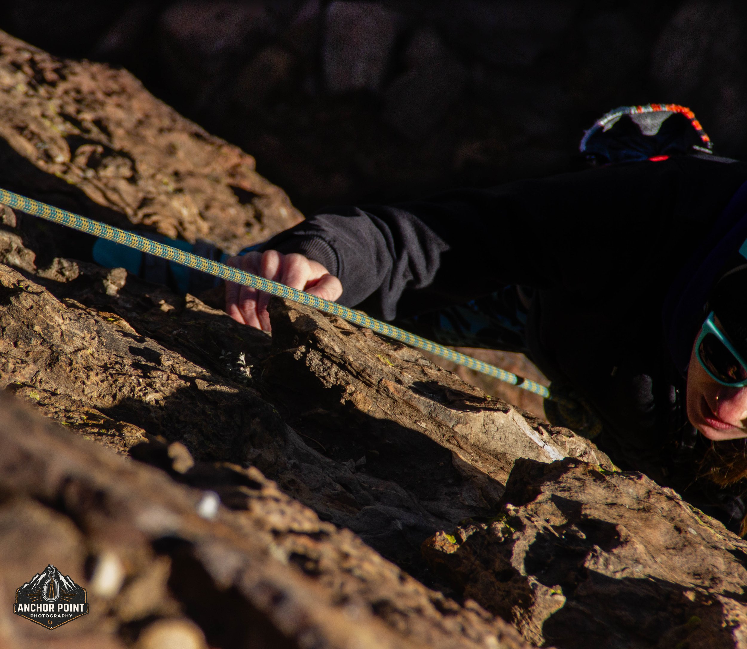 A person rock climbing on a rugged cliff face, wearing sunglasses and a black long-sleeve shirt, gripping the rock with their right hand and secured with a yellow climbing rope.