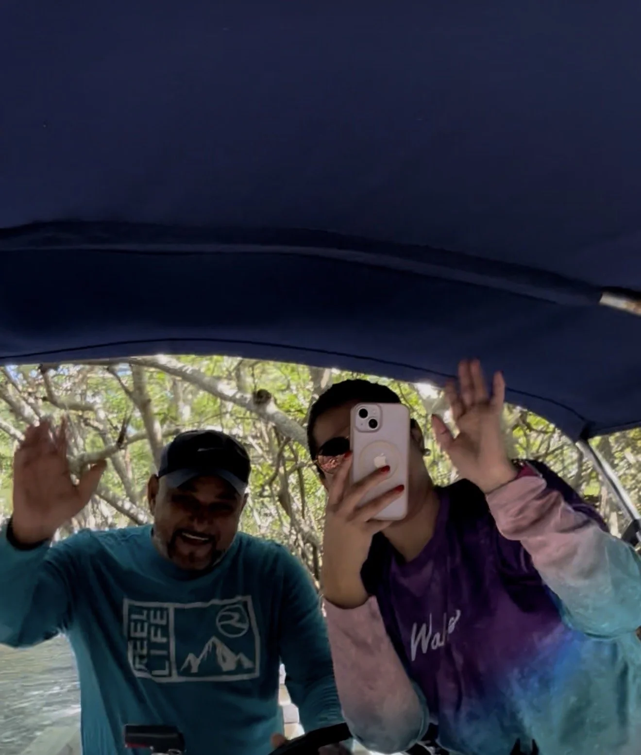 Two people taking a selfie outdoors under a canopy of trees, both smiling and waving, one holding a smartphone with a pink case.